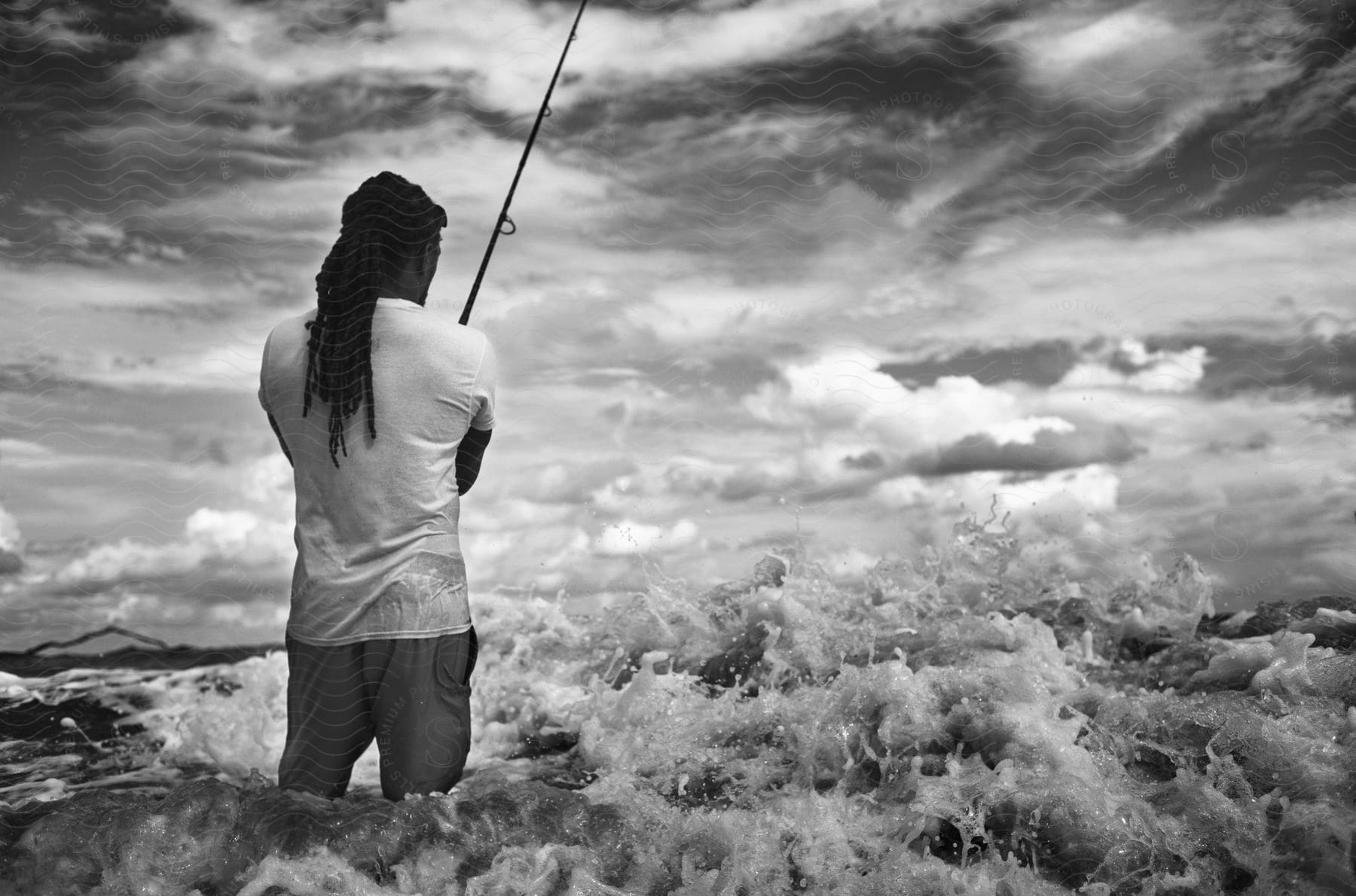 Man with his back and inside the beach with the sea waves holding a fishing rod on a sky day with clouds.