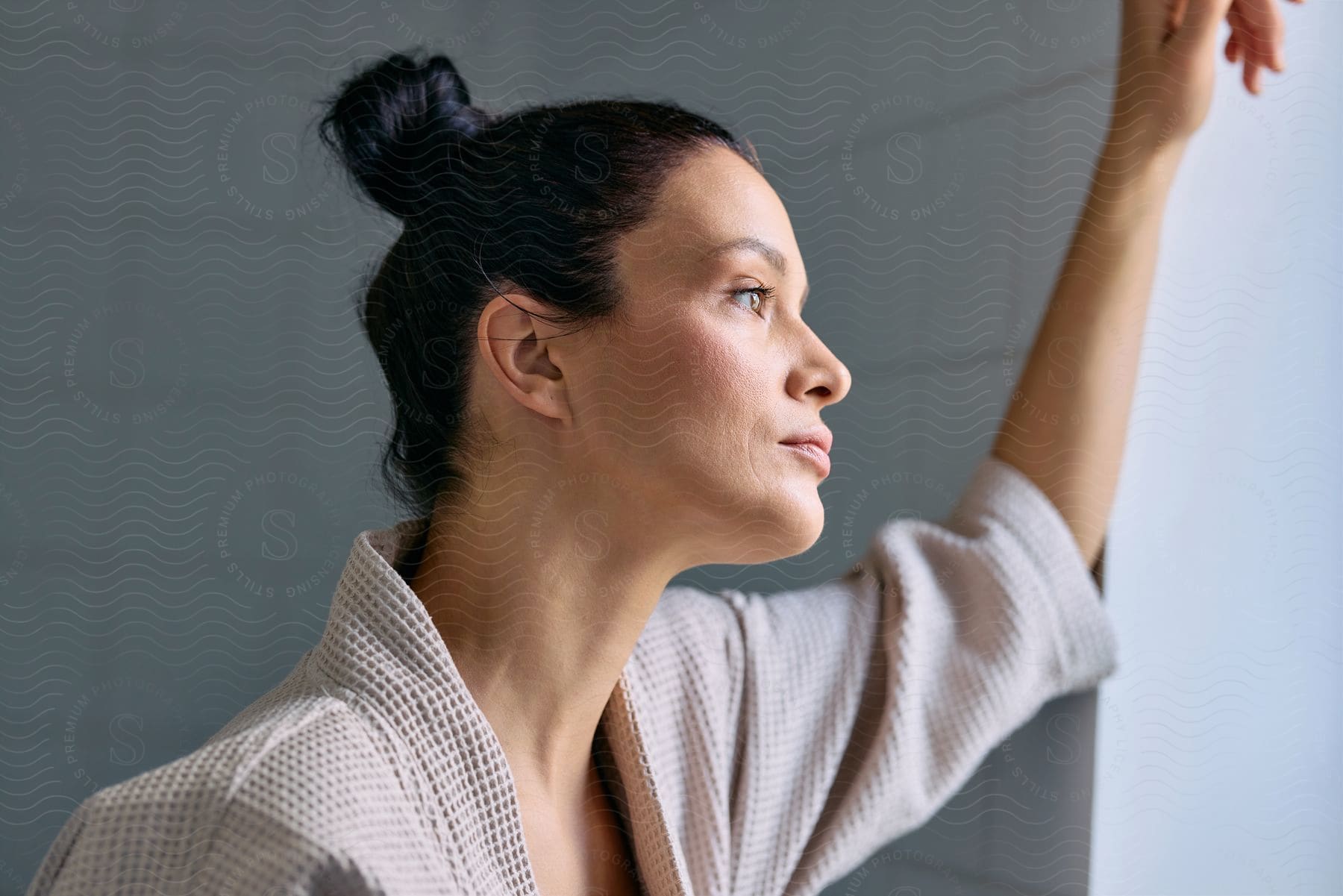 Woman wearing blush and a robe leans against bathroom wall while looking out the window.