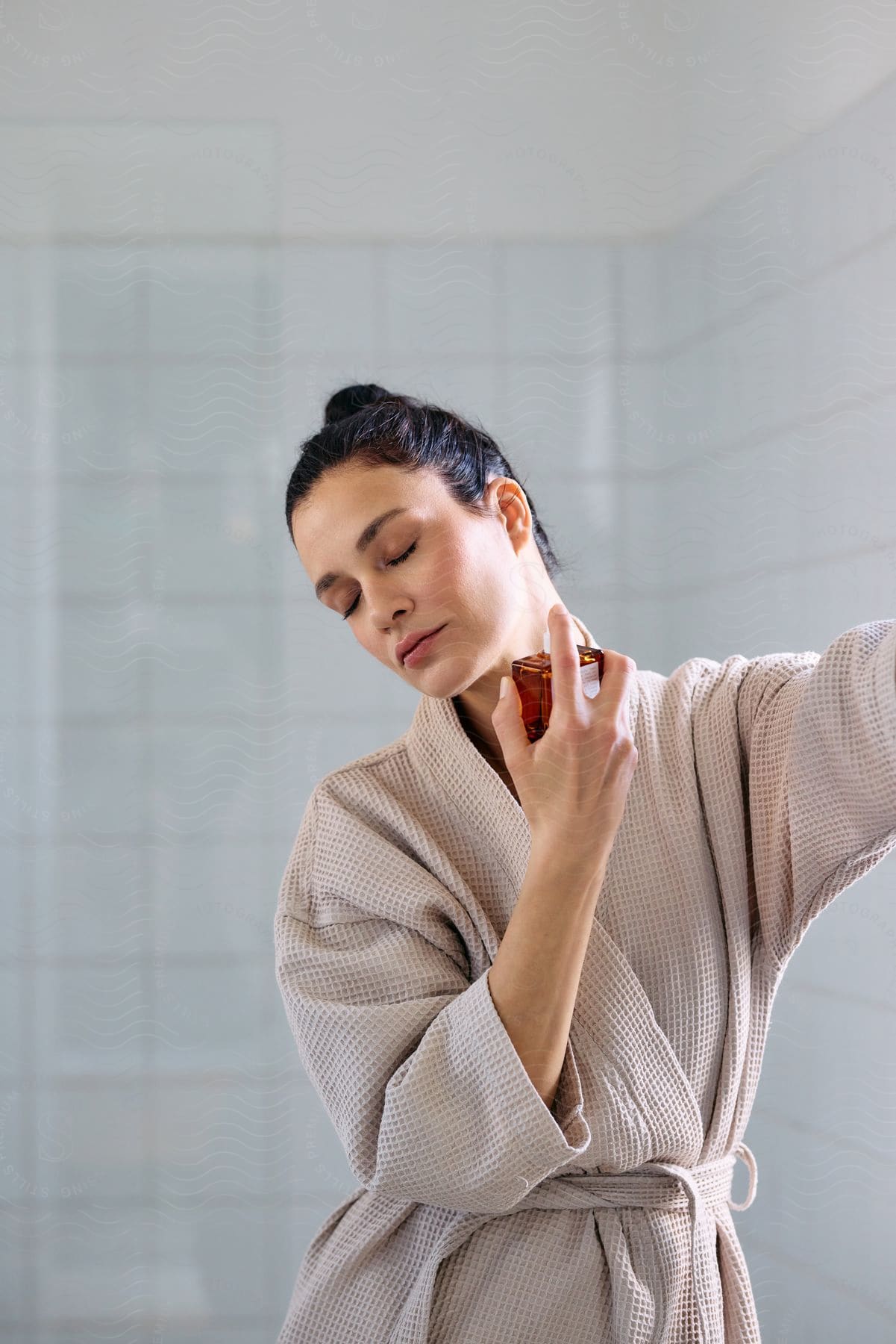 Woman wearing a robe while standing in the bathroom sprays perfume onto her neck.