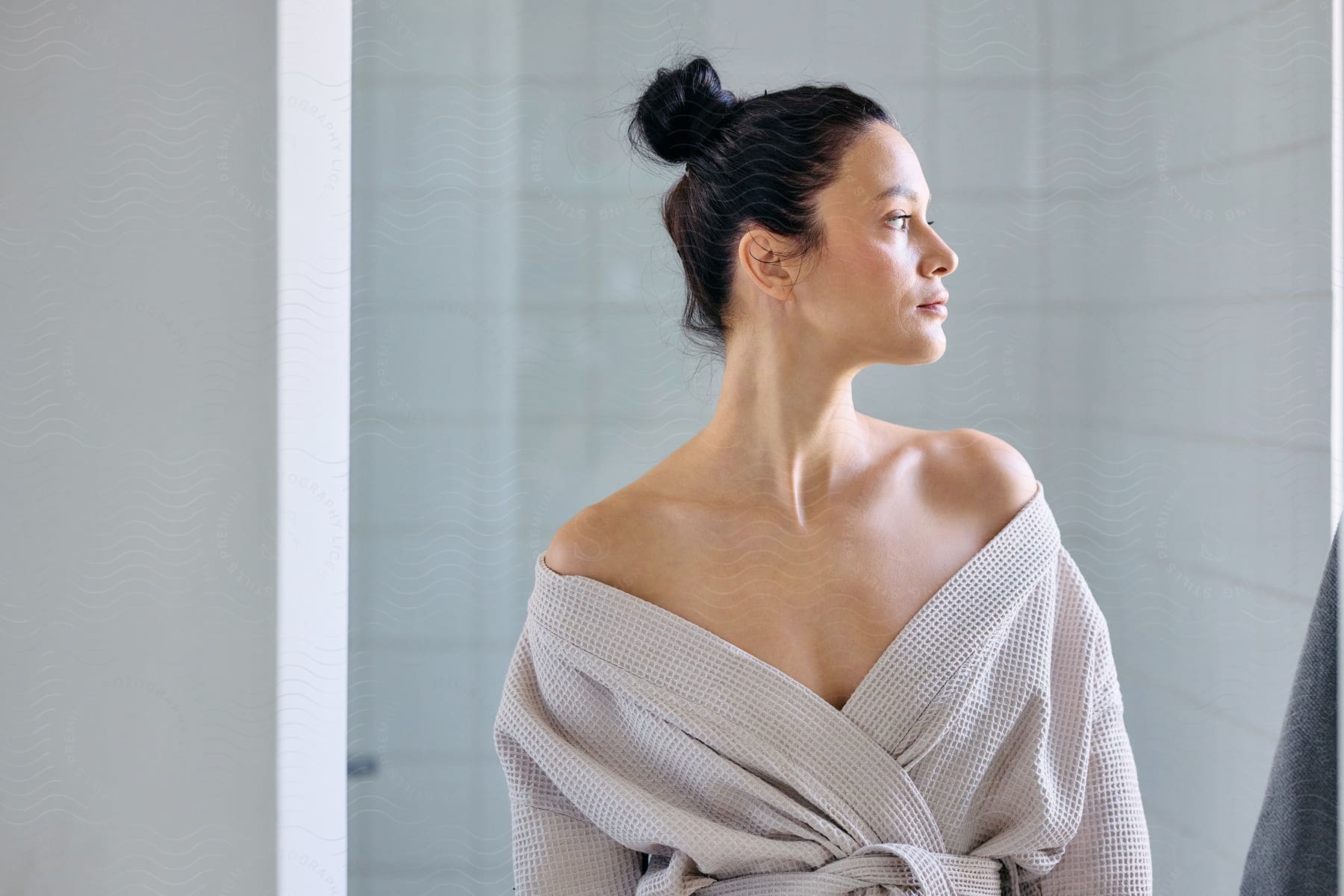 A woman in a bathroom, looking to her side, with her bath robe lowered to her shoulders.
