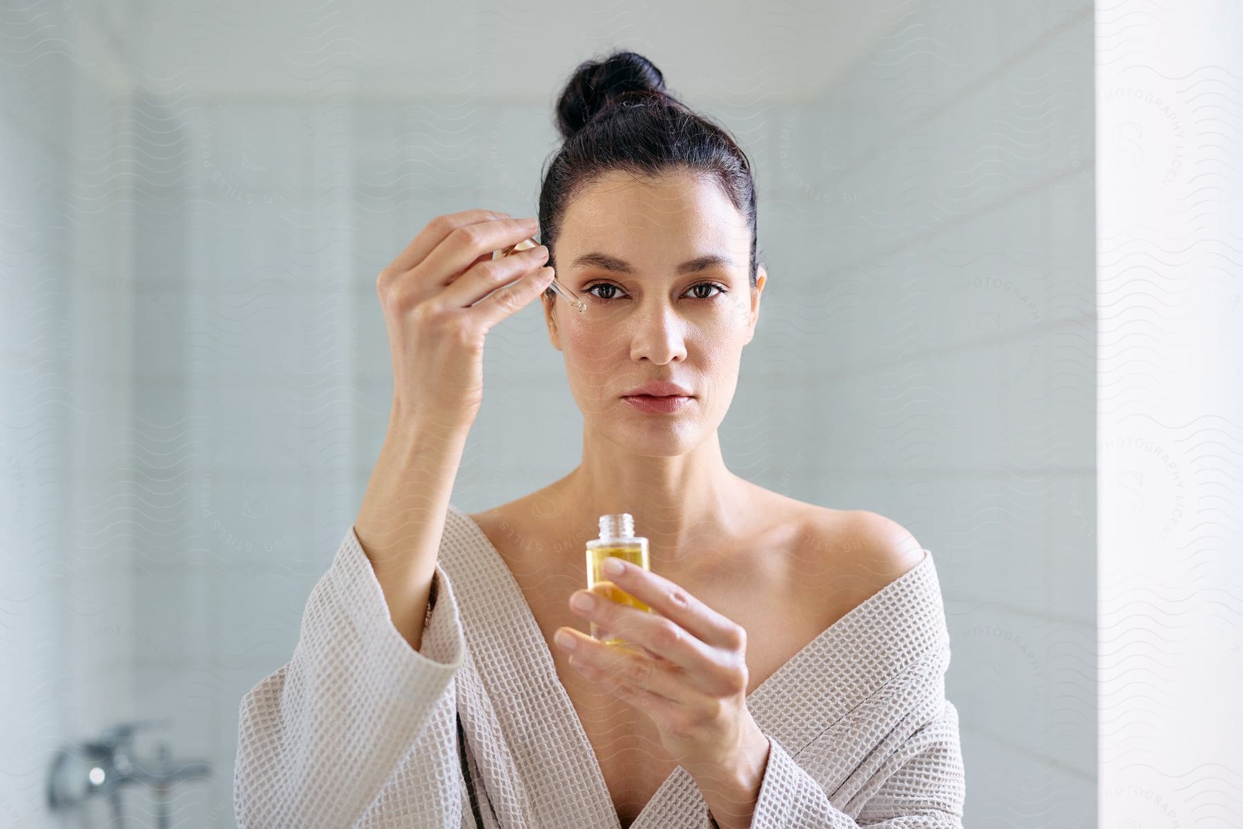 Woman holding a dropper of serum over its bottle in a white bathroom.