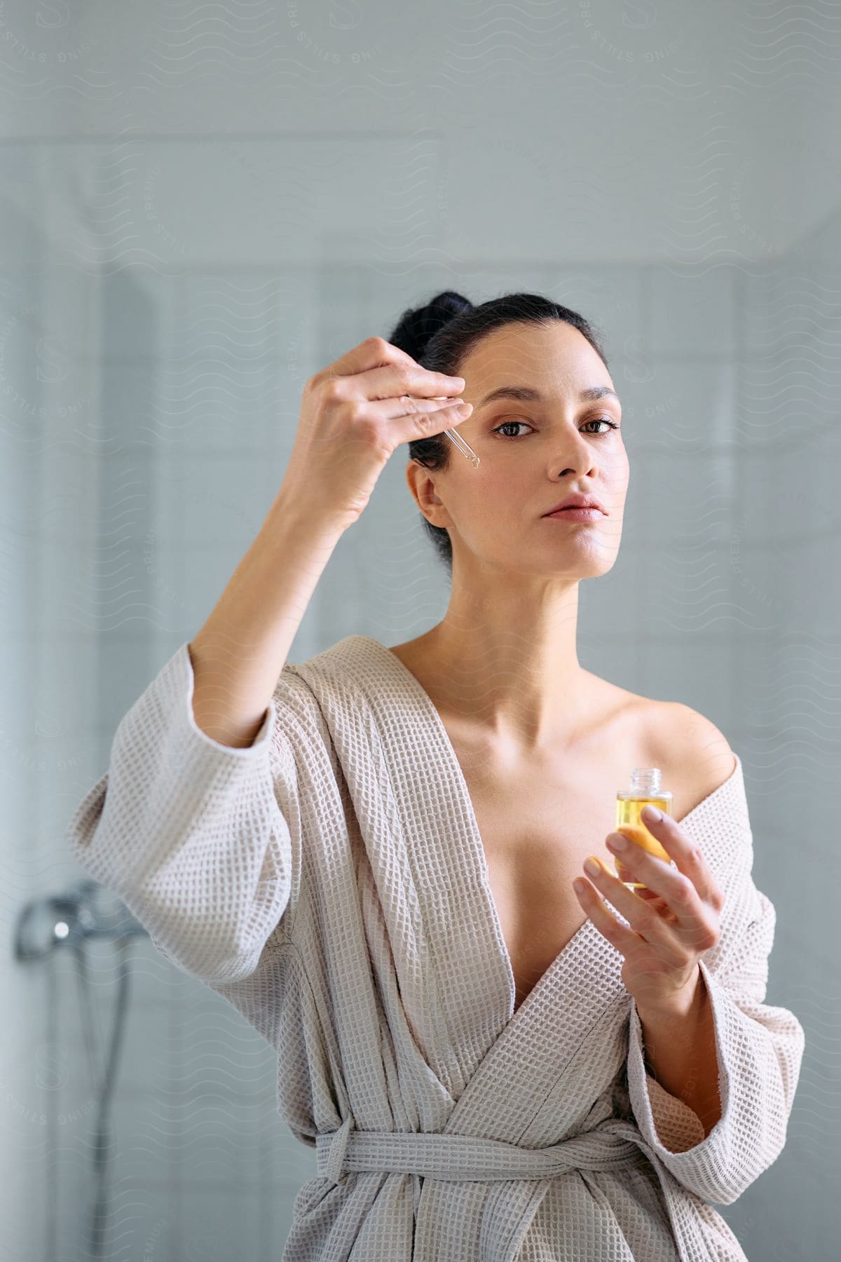 A woman in a bathrobe is applying a product to her face in a bathroom.