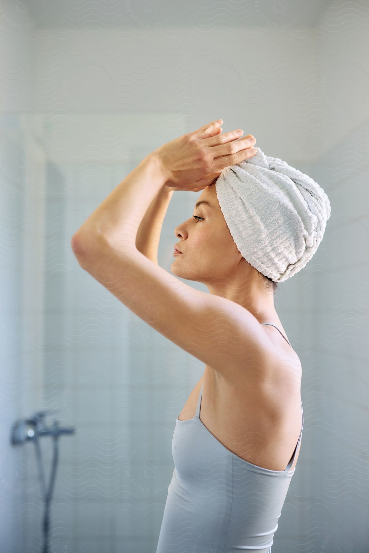 A woman stands in the bathroom with her head wrapped in a towel.