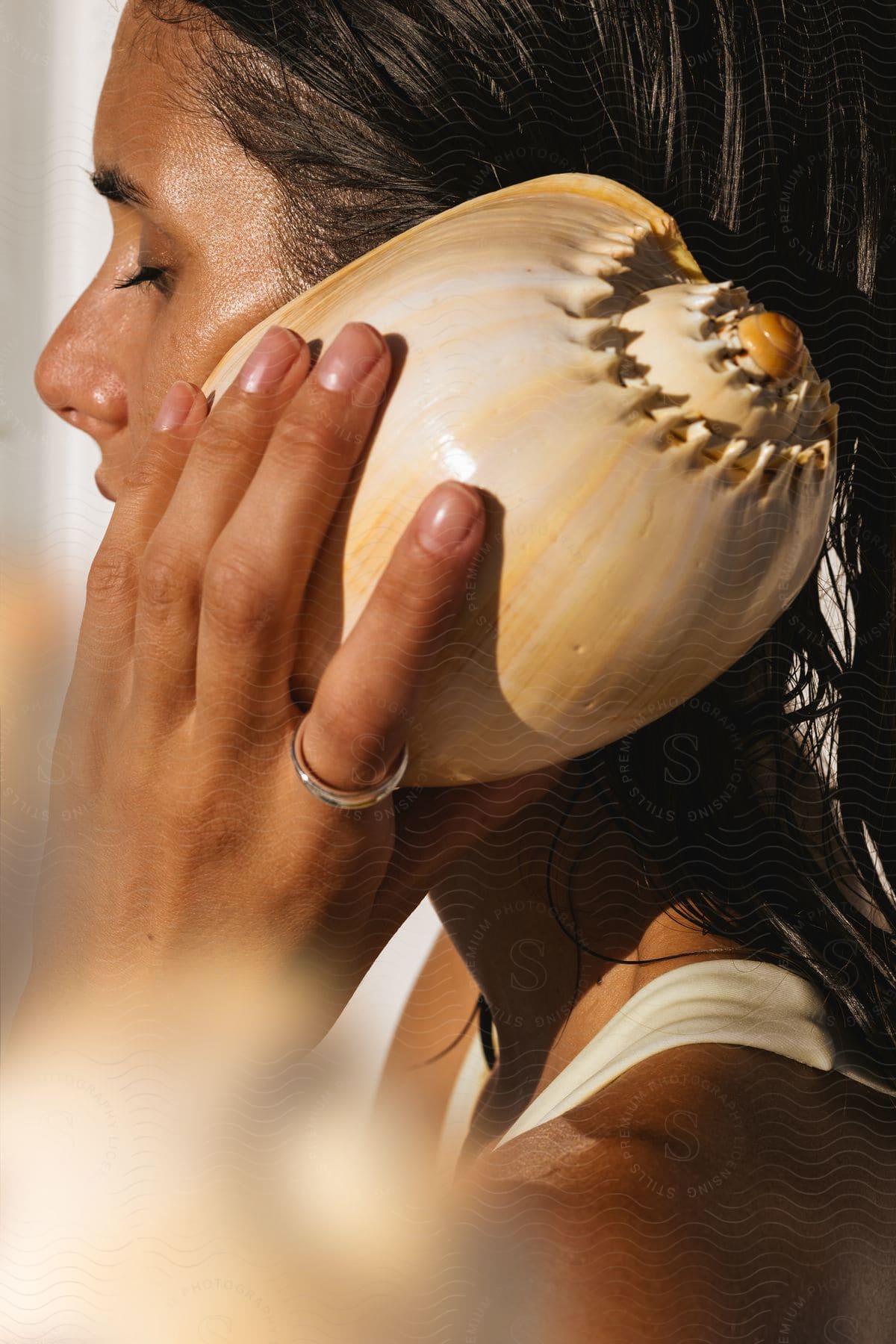 A brunette woman puts a huge shell in her ear and is closing her eye and her skin is reflecting natural light