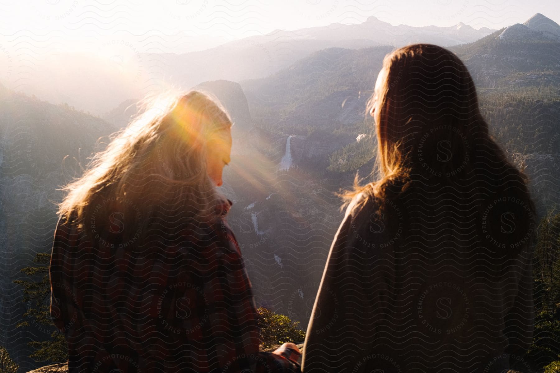 Two women with their backs are at a high point observing a landscape of a valley between some mountains and the women are being illuminated by the sunlight