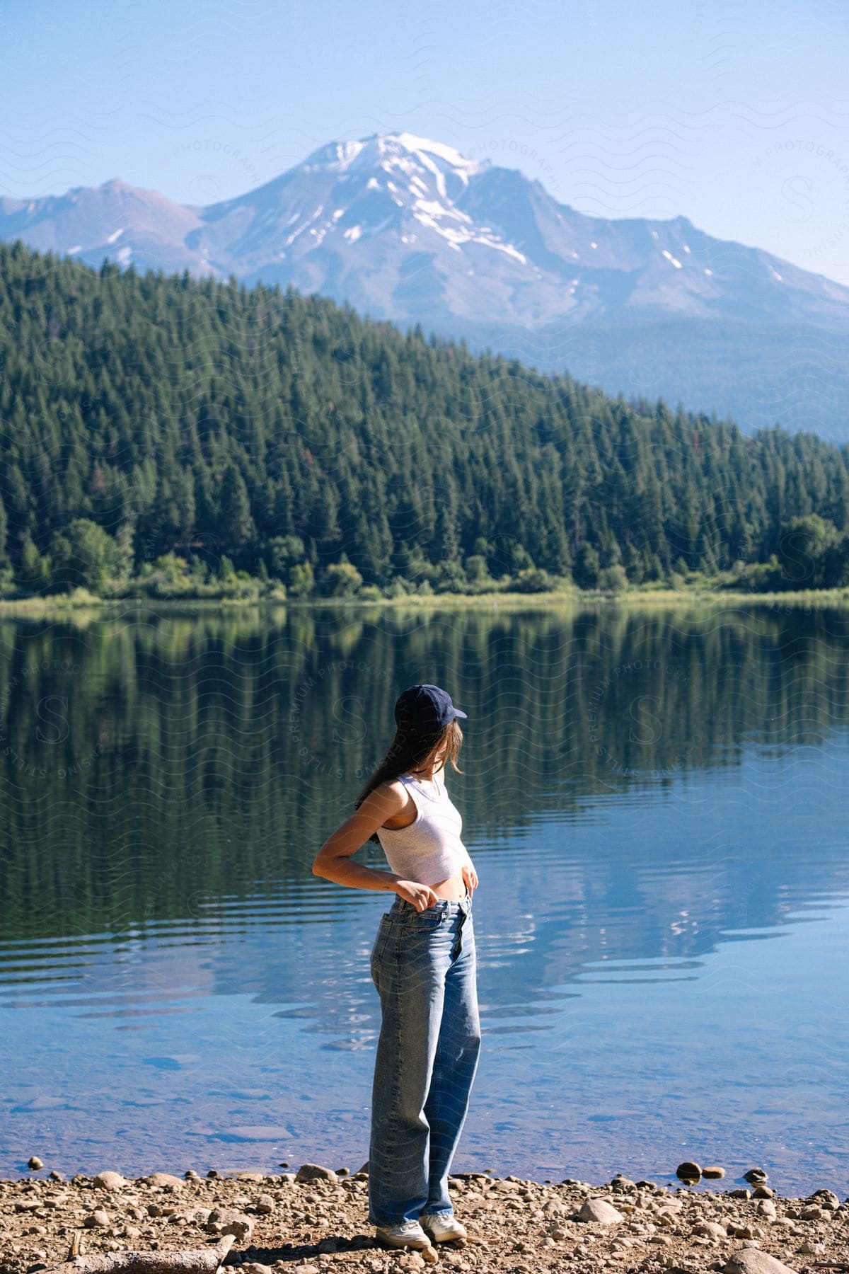 Woman standing by a lake with a view of a forested hillside and snow-capped mountain in the background.