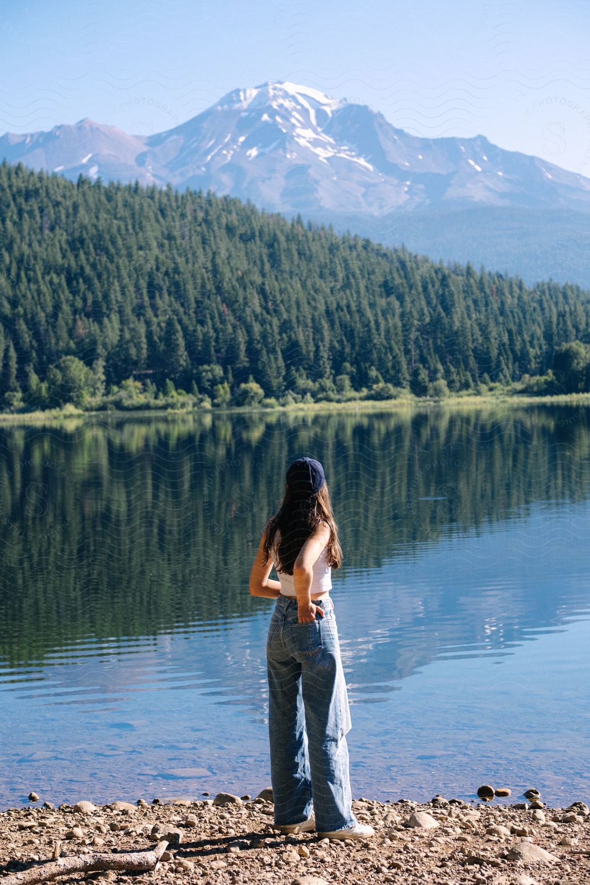 On a sunny day, a woman wearing jeans, sneakers, and a hat stands on the river bank, gazing at the river. Tall trees surround her on one side, with a mountain in the backdrop.