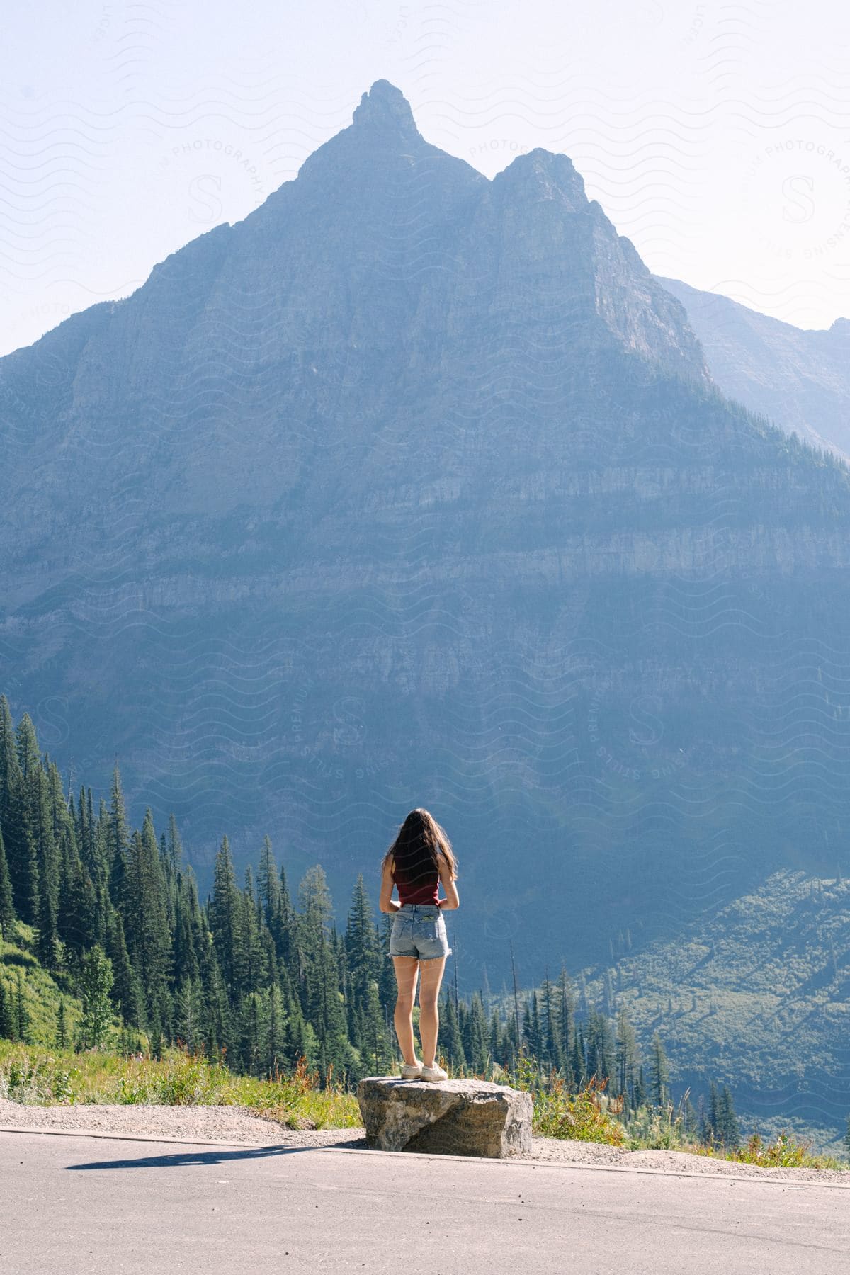 Woman standing on a rock on the side of the road overlooking a large mountain with cliffs during the day