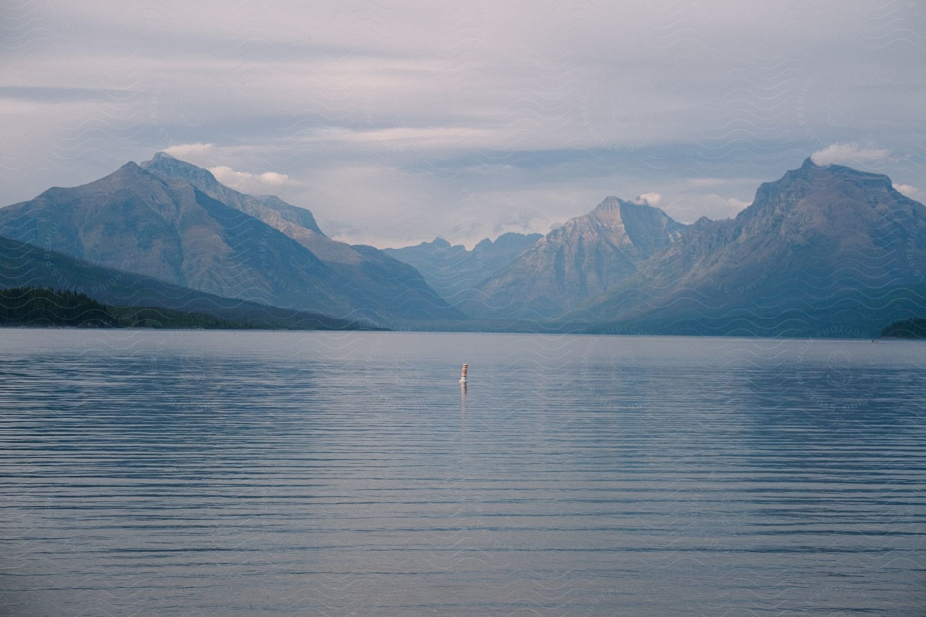 Lake with a single buoy and mountain range in the distance under an overcast sky.