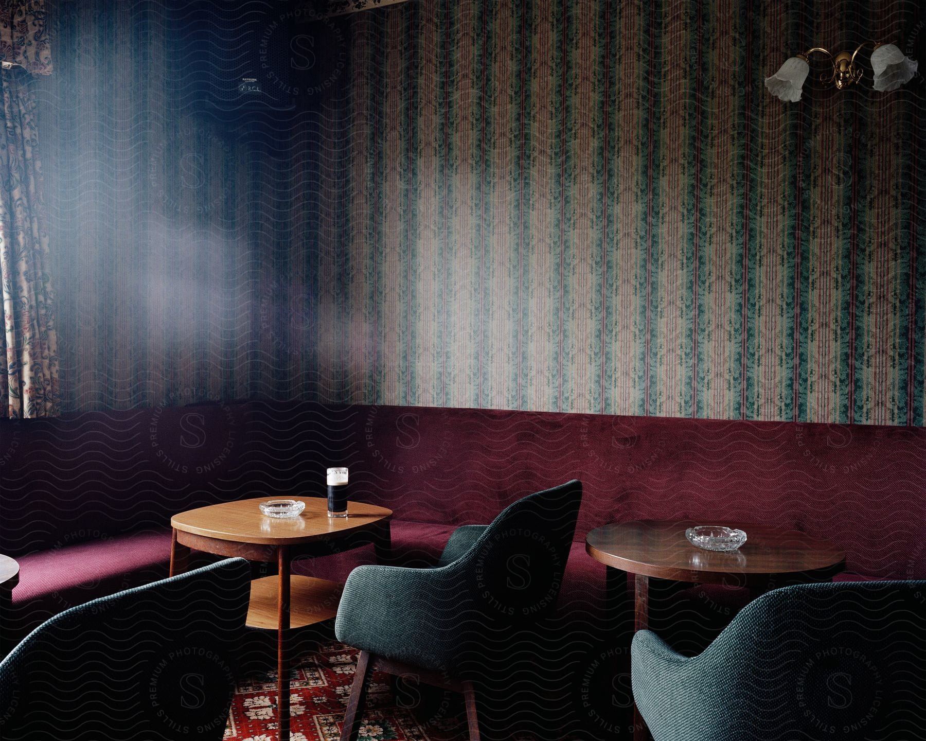 The interior of an establishment where it is possible to see a red bench bordering the walls accompanied by two wooden tables with some chairs on the table, crystal ashtrays and a glass with a drink