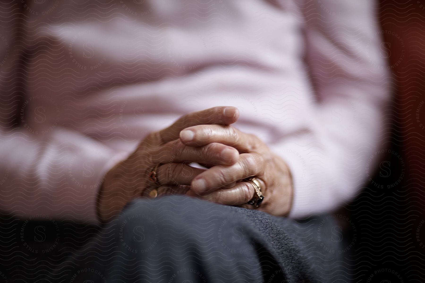 Close-up of clasped hands with rings on the fingers, resting on a lap with a pink shirt and gray pants in the background.