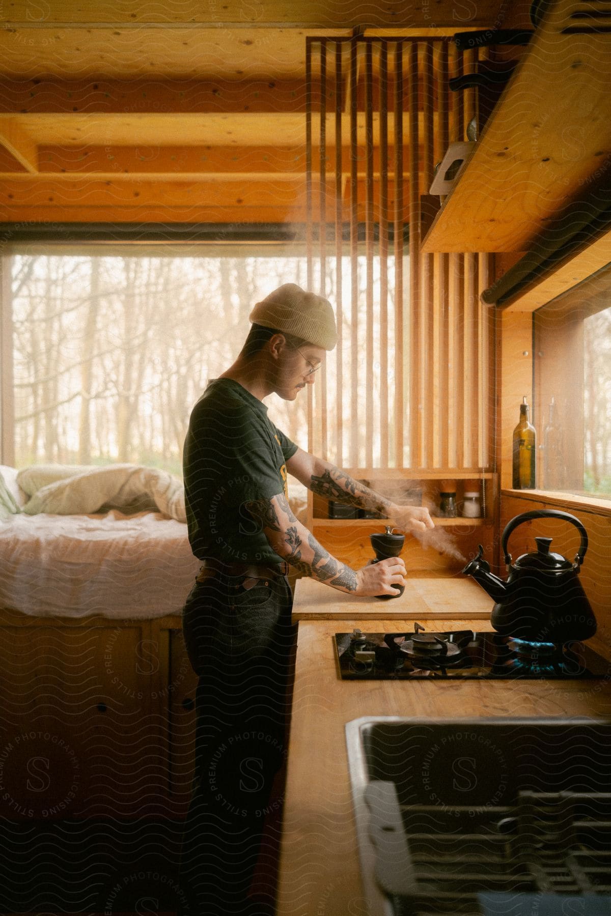 A man with a mustache and tattoos on his arms, wearing a white wool hat, T-shirt, jeans, and a silver wristwatch, grinds coffee beans on a wooden counter in the kitchen.