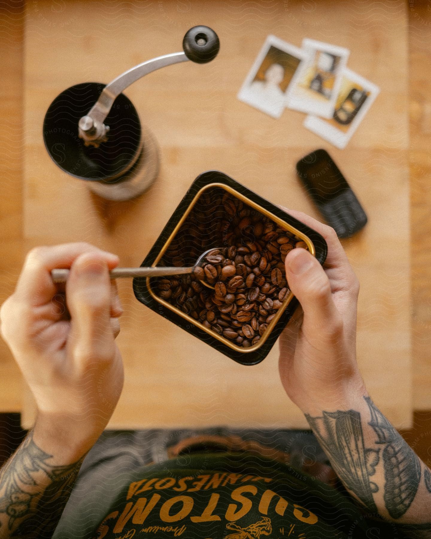 A person with tattooed hands is seen holding a plate of raw coffee beans and serving them into a hand grinder using a spoon. The grinder is placed on a table, with a mobile device also resting on the table.