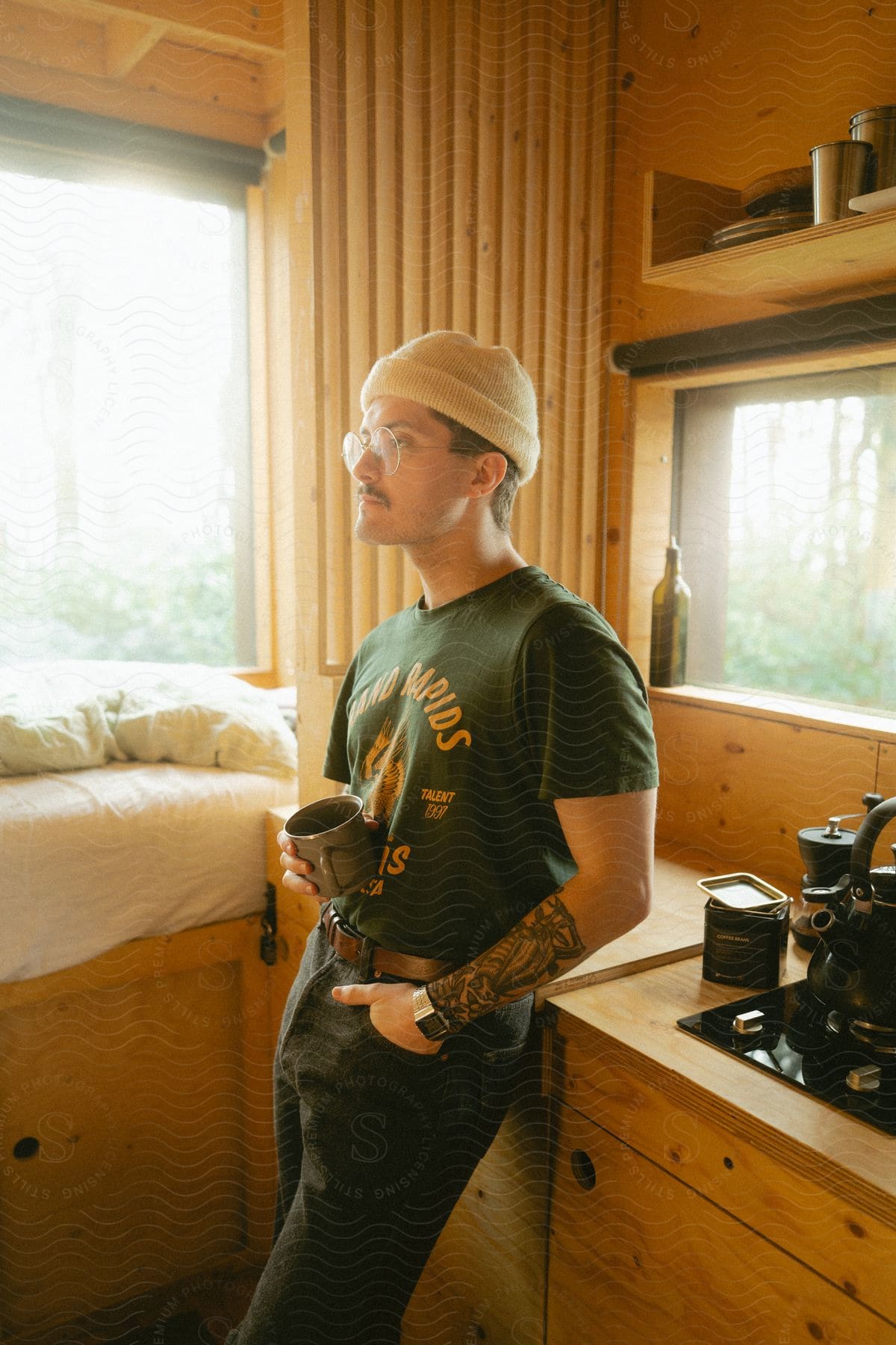 A man with a mustache and tattoo on his left arm, wearing a white wool hat, T-shirt, jeans, and a silver wristwatch, leans against a wooden kitchen counter holding a cup and looking into space.