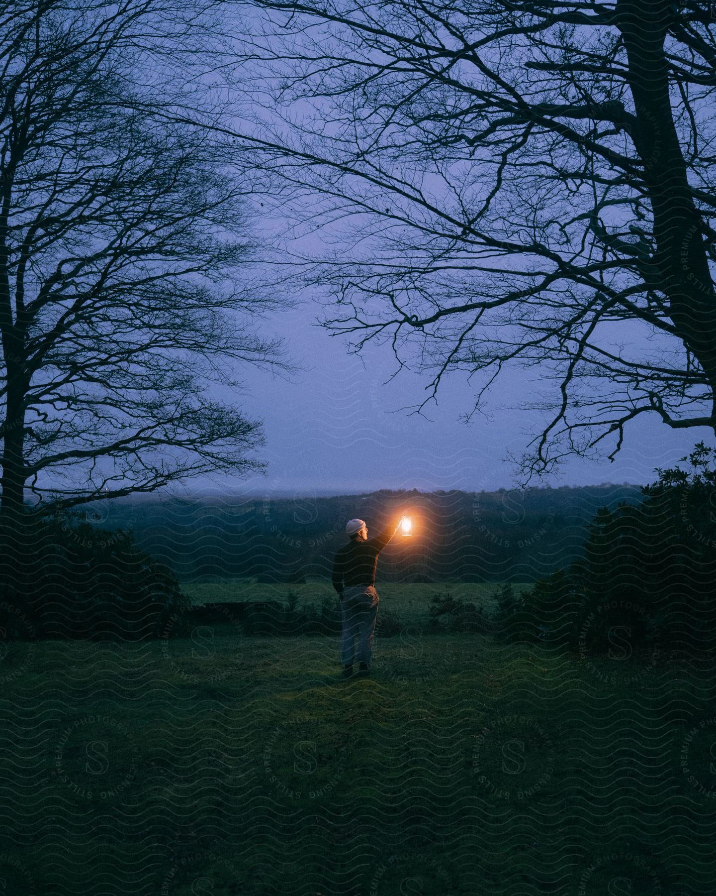 A man holds a lantern up while standing outside at dusk.