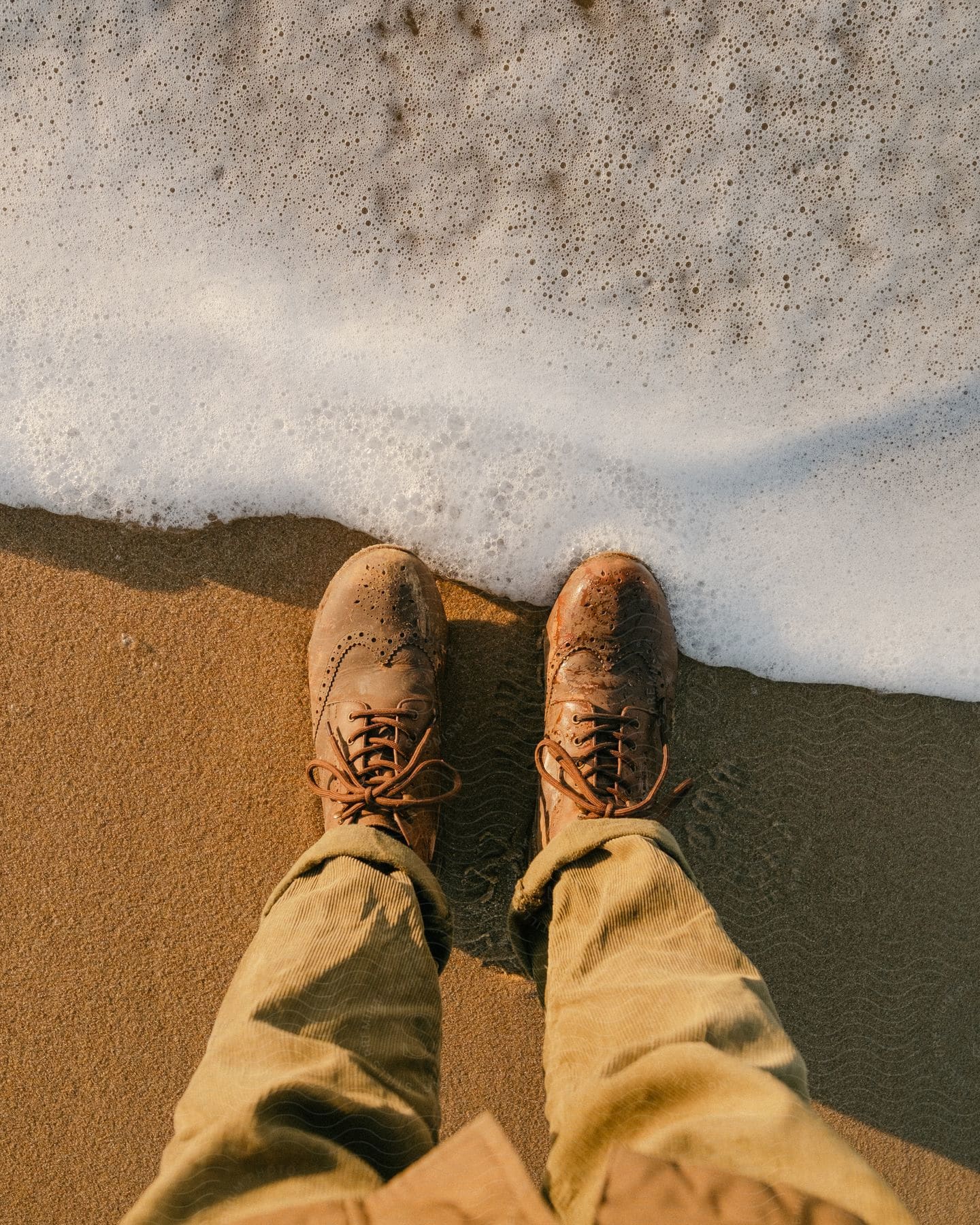 Legs and feet of a person in leather boots in the sand and at the edge of the foamy sea water.