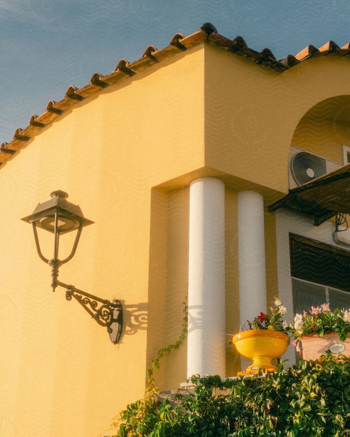 Exterior architecture of a house with a yellow wall, a vintage chandelier and vases with flowers next to it.