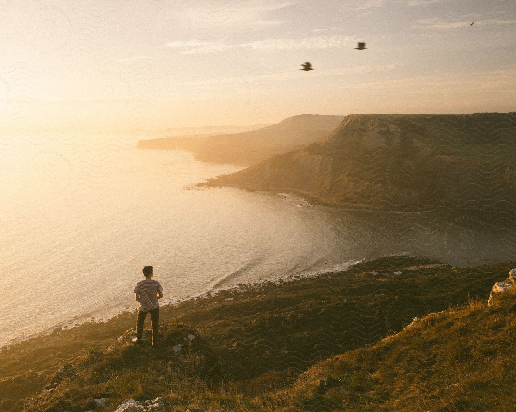 Man wearing white t shirt and jeans stands on a rock watching the coastline at sunset