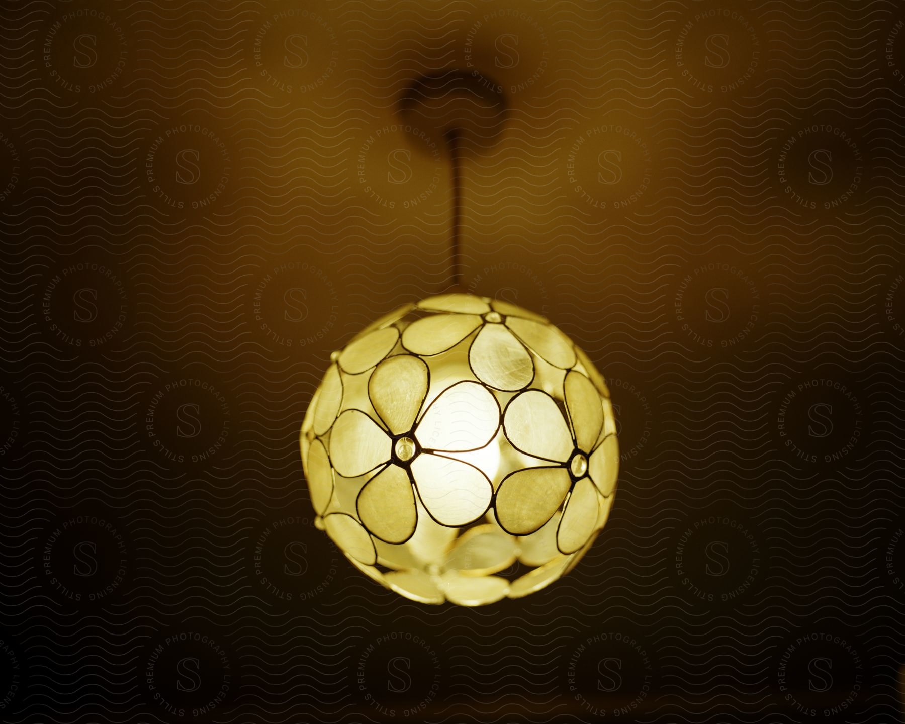 A decorative chandelier with a floral pattern, emitting a soft light in a dark environment. The translucent petals form a flower-like design, creating an ambient lighting effect.