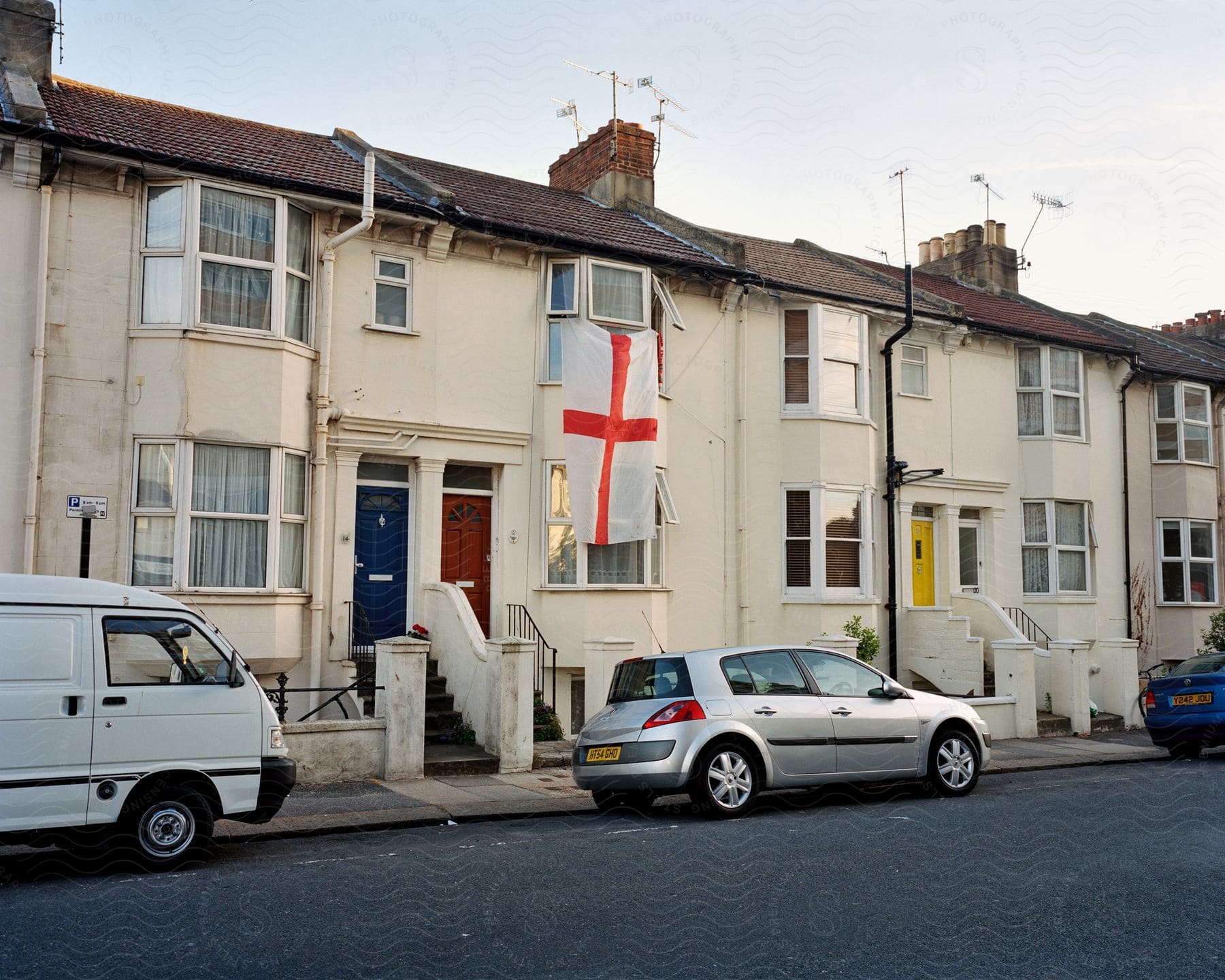 A row of houses along a street, with a large English flag hanging from one of them. The houses are old, with signs of wear and tear, and there are three cars parked in the street in front.