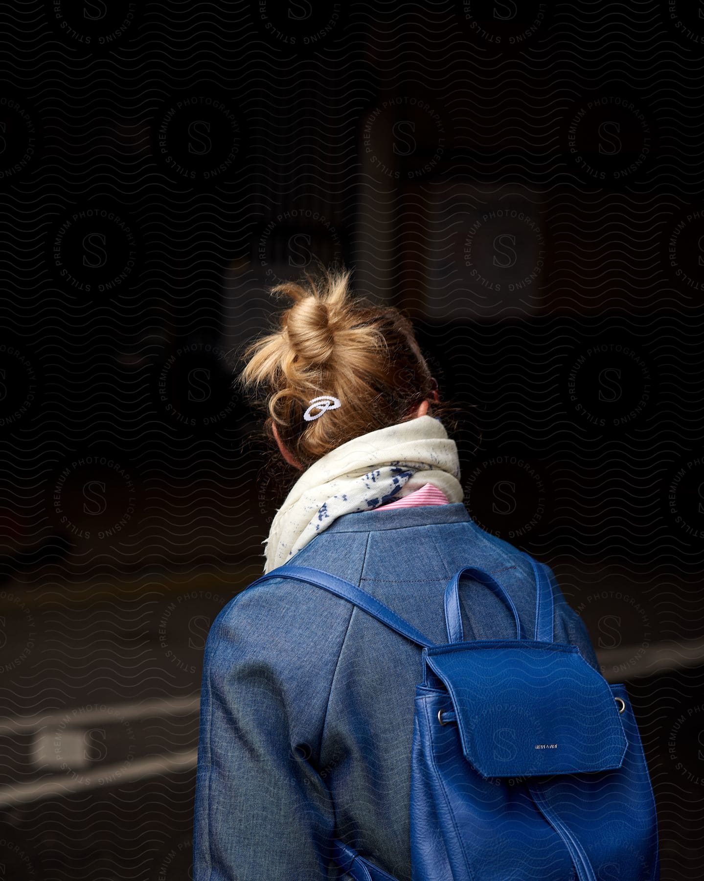 A blonde woman wearing a blue coat and backpack looks sideways.