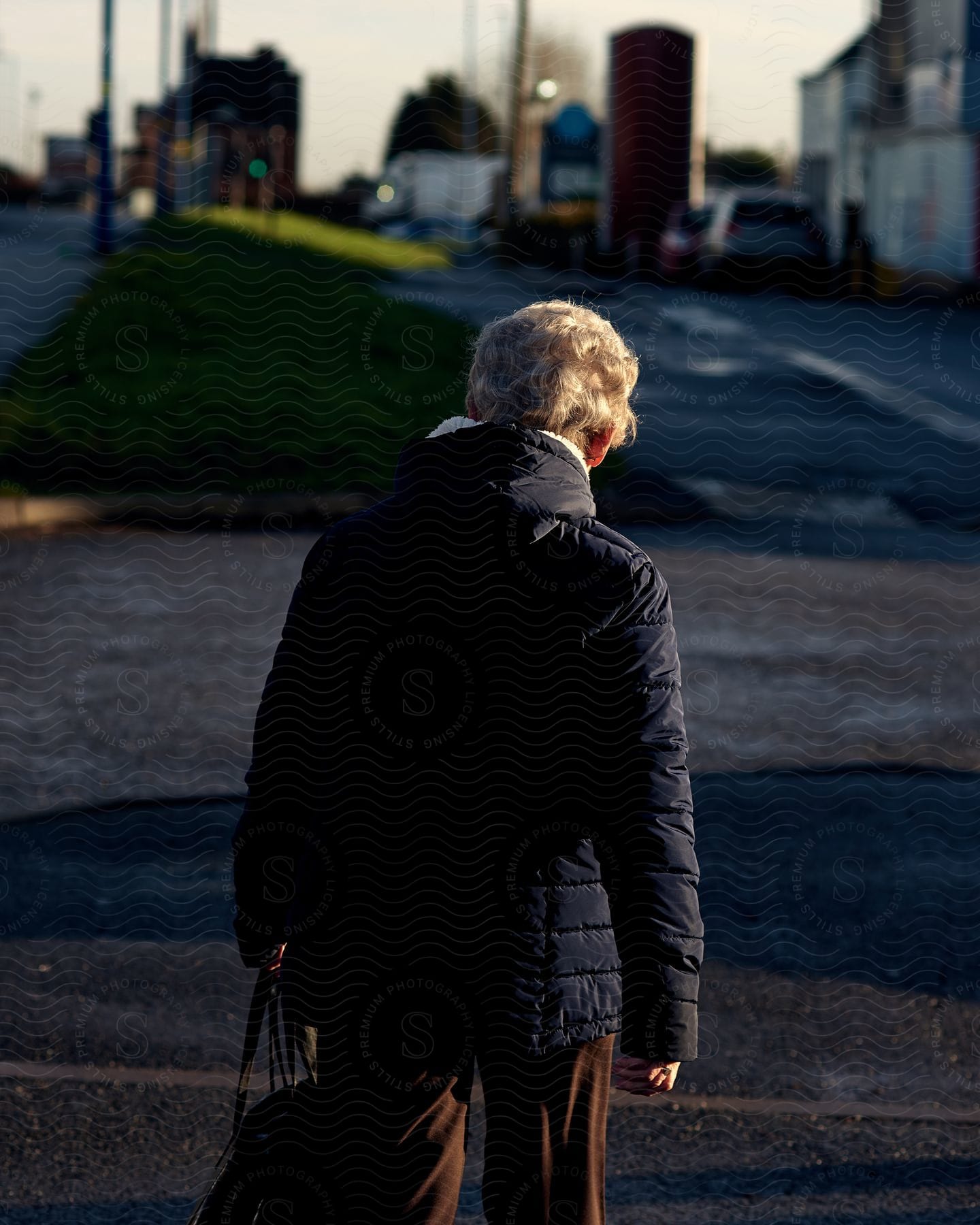 Elderly person with his back turned walking down a street wearing a long-sleeved jacket and holding a bag.