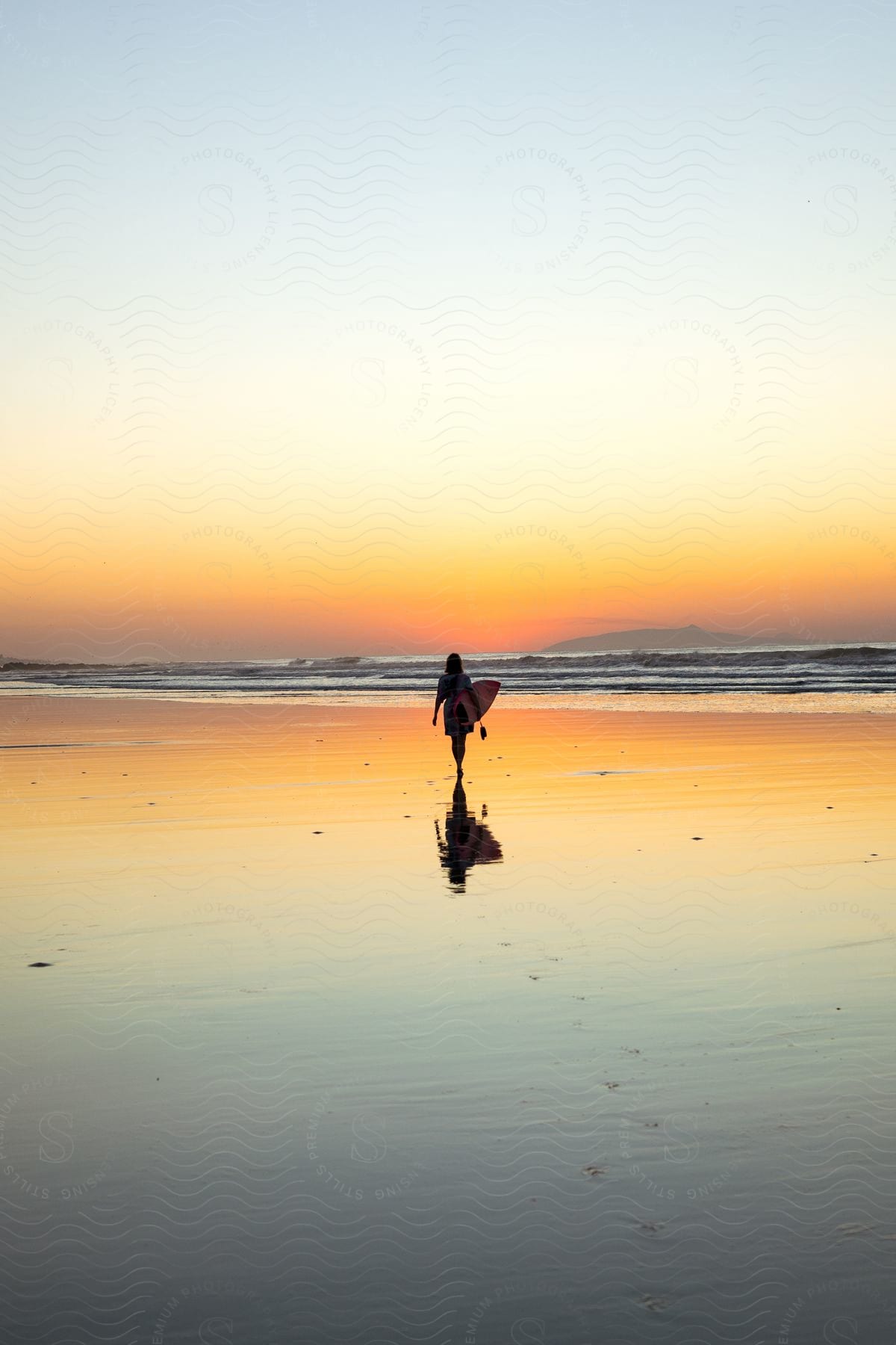 The silhouette of a woman carrying a surfboard is reflected on the golden beach as she walks toward the waves