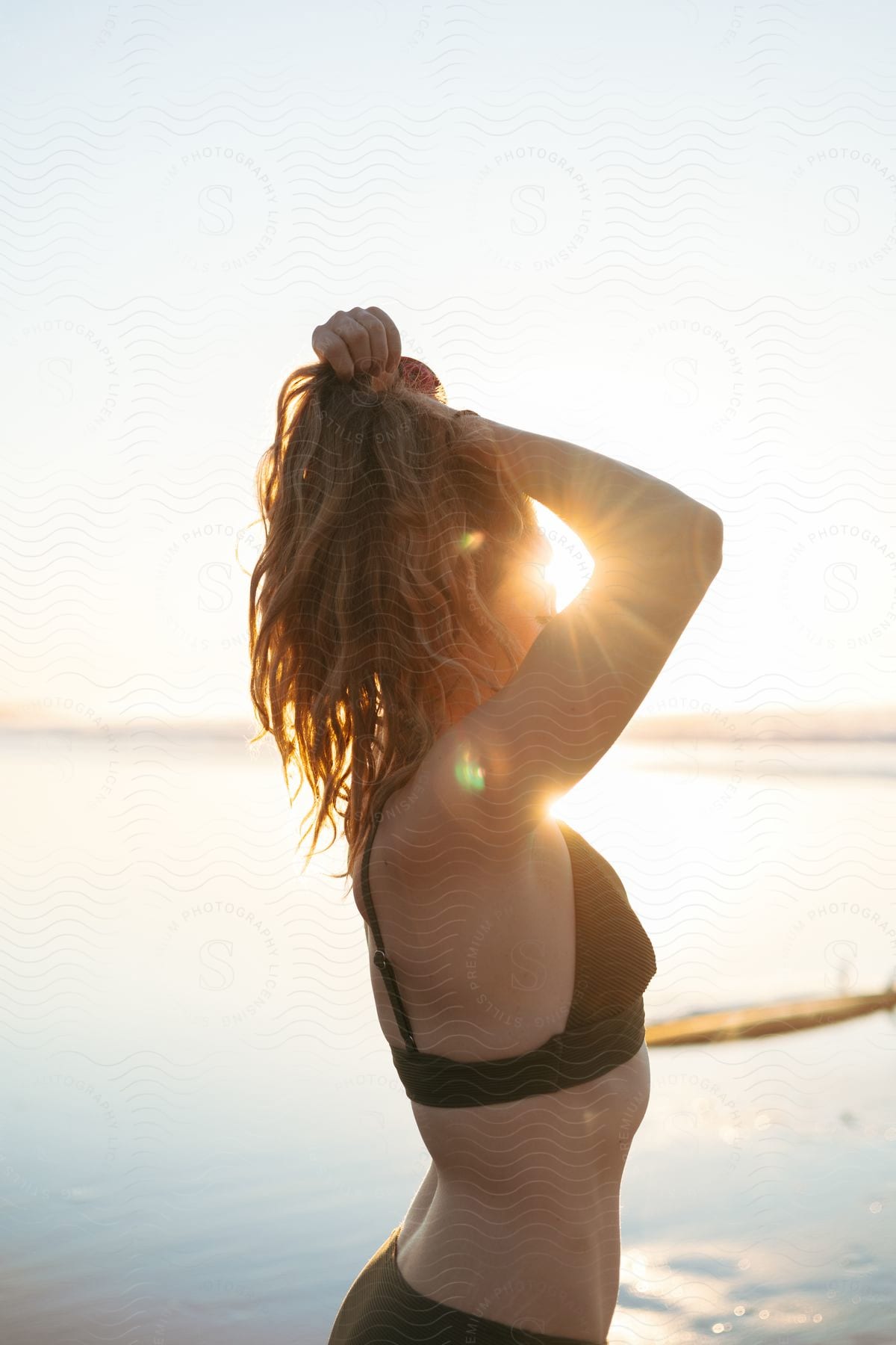 A woman in a bikini standing above the water with arms up and hands in her hair as the setting sun shines on her