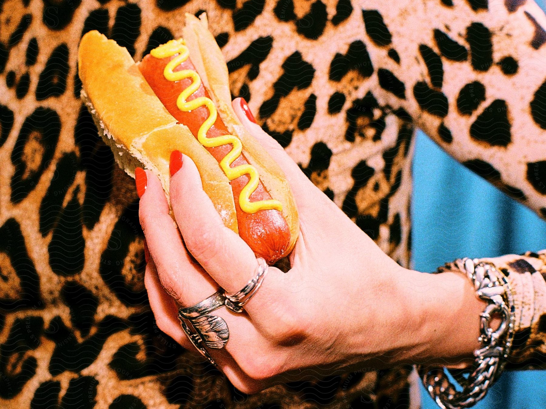 Woman hand adorned with silver rings and bracelets holds a hot dog with mustard.