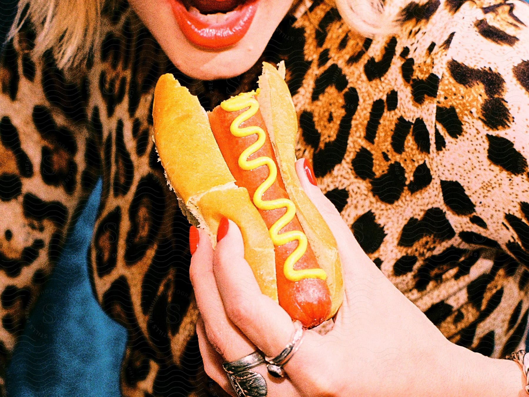Woman brings an already-bitten hotdog with mustard on it closer to her open mouth with her left hand.