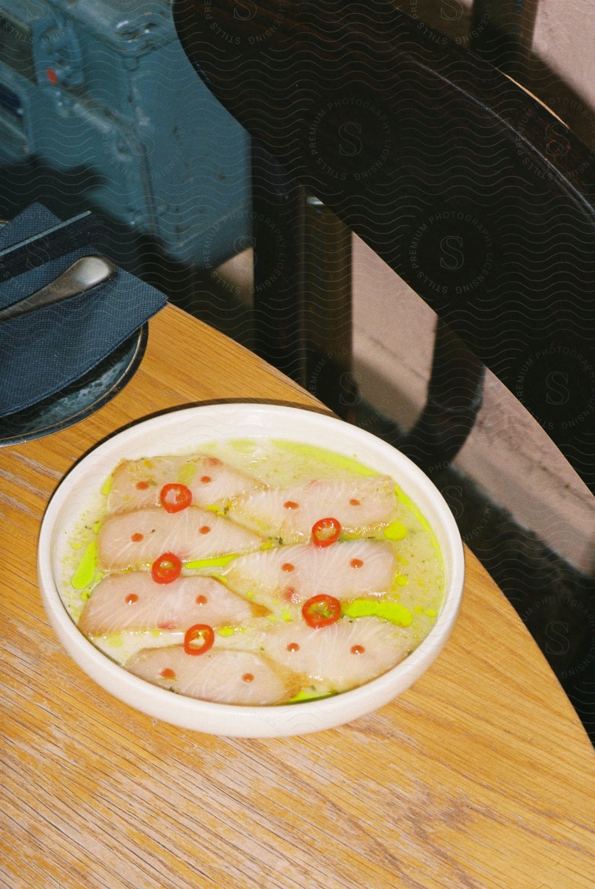 A bowl of food with slices of a light-colored ingredient topped with small red items and a green sauce and plate is on a wooden table