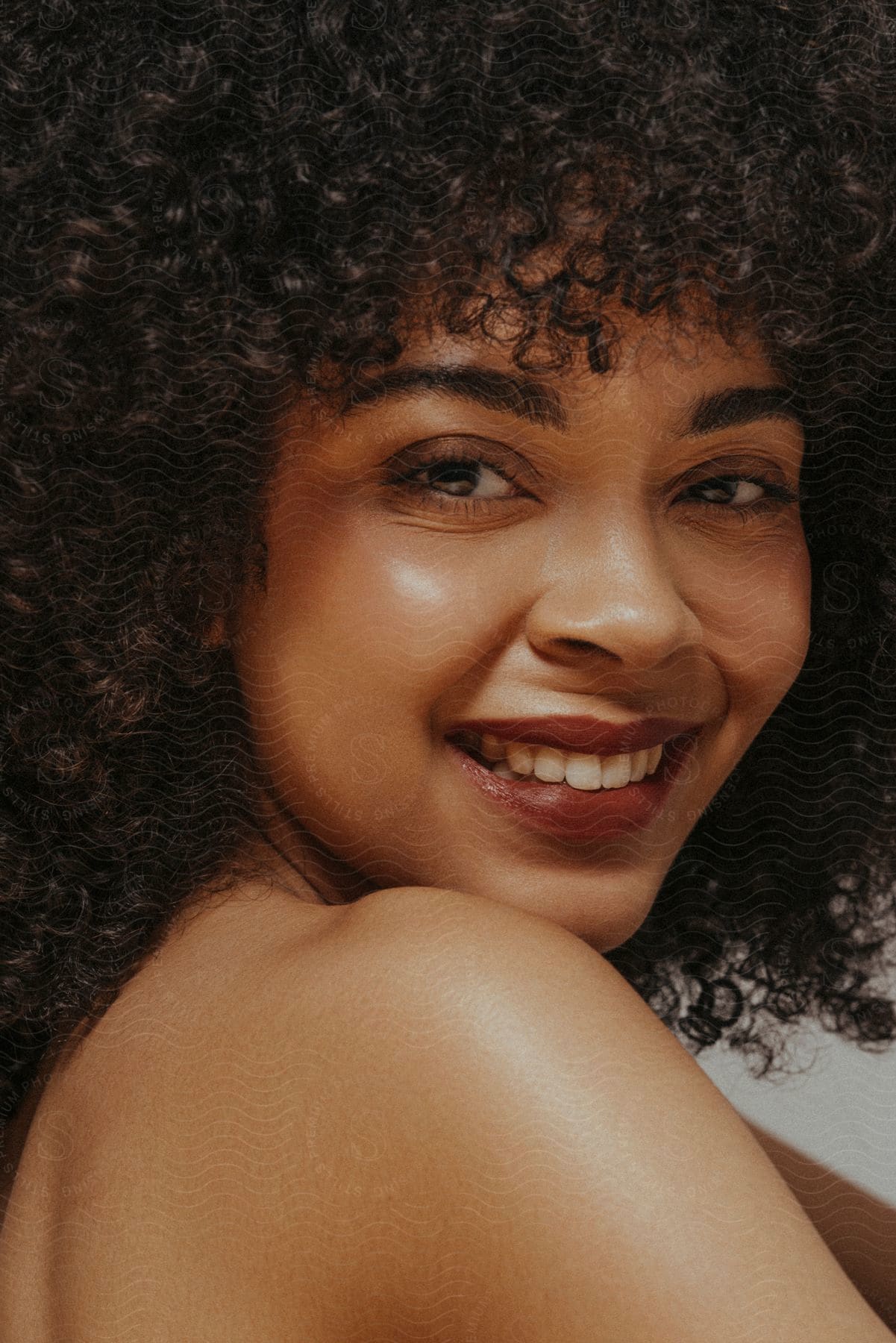 Woman with curly hair smiling over her shoulder, bare shoulders visible.