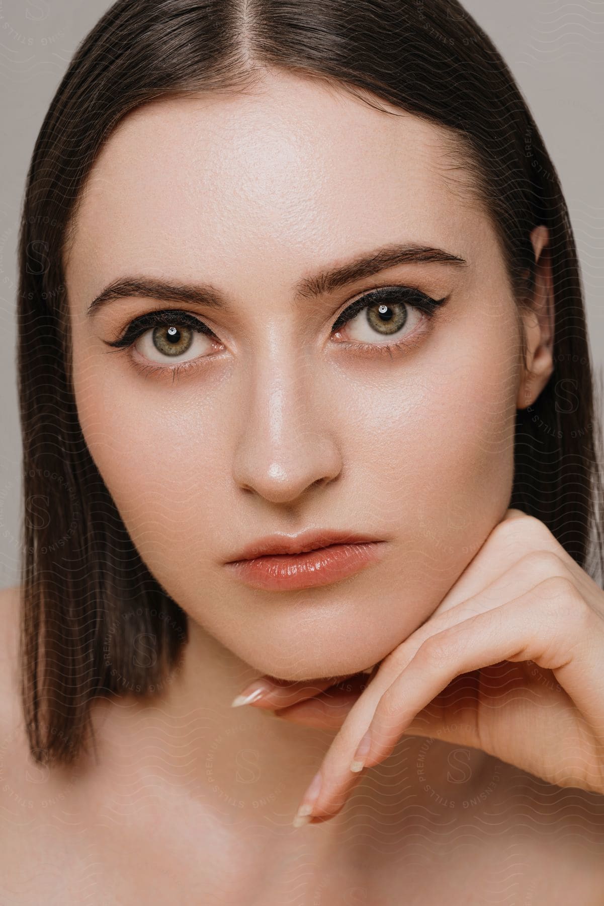 A close-up portrait of a woman with dark hair, neutral makeup, and her hand resting gently against her chin.