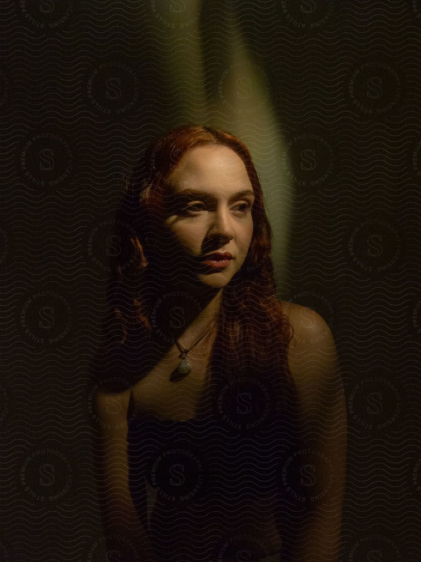 In a dimly lit room, a white woman with red hair wears a necklace.