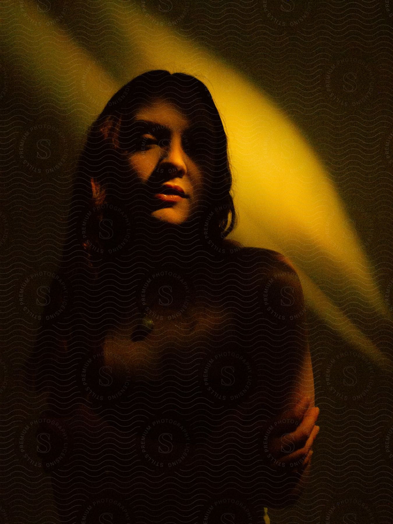 Woman in shadow with crossed arms, face illuminated by a slanted beam of light, against a dark backdrop.