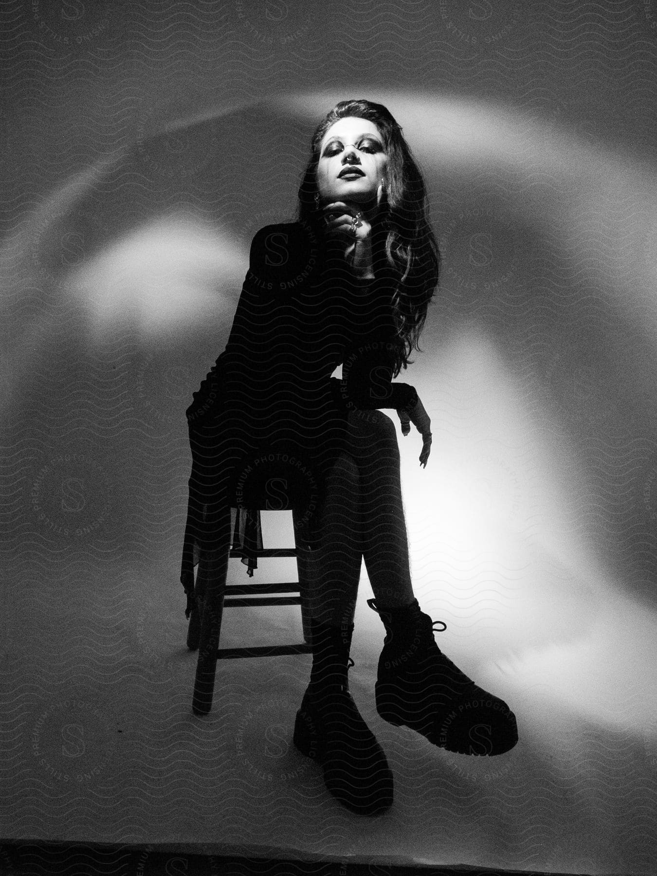 Woman posing sitting on a wooden stool with her legs crossed and a hand on her chin on a white studio background.