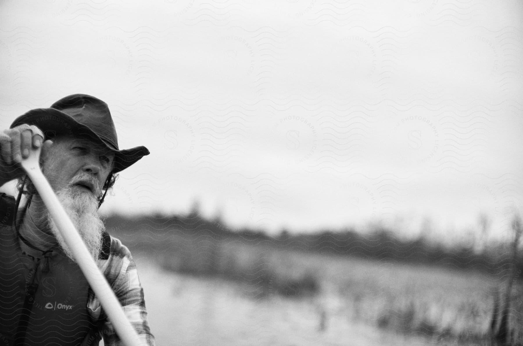 On a sunny day, an elderly man with long beard wearing a hat and holding a stick on a sunny day.