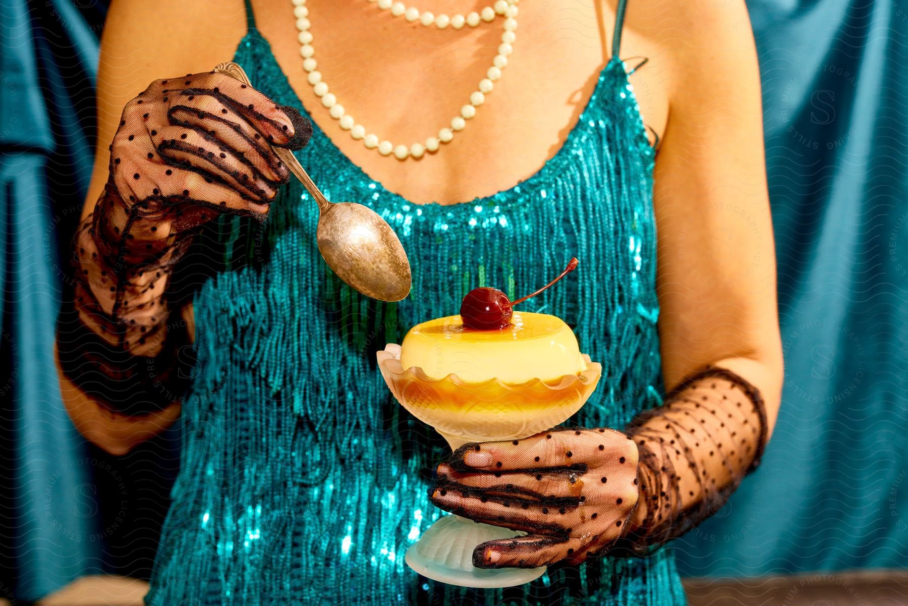 Woman in blue dress and pearl necklace with decorated black gloves holding a dessert cup with a cherry on top.