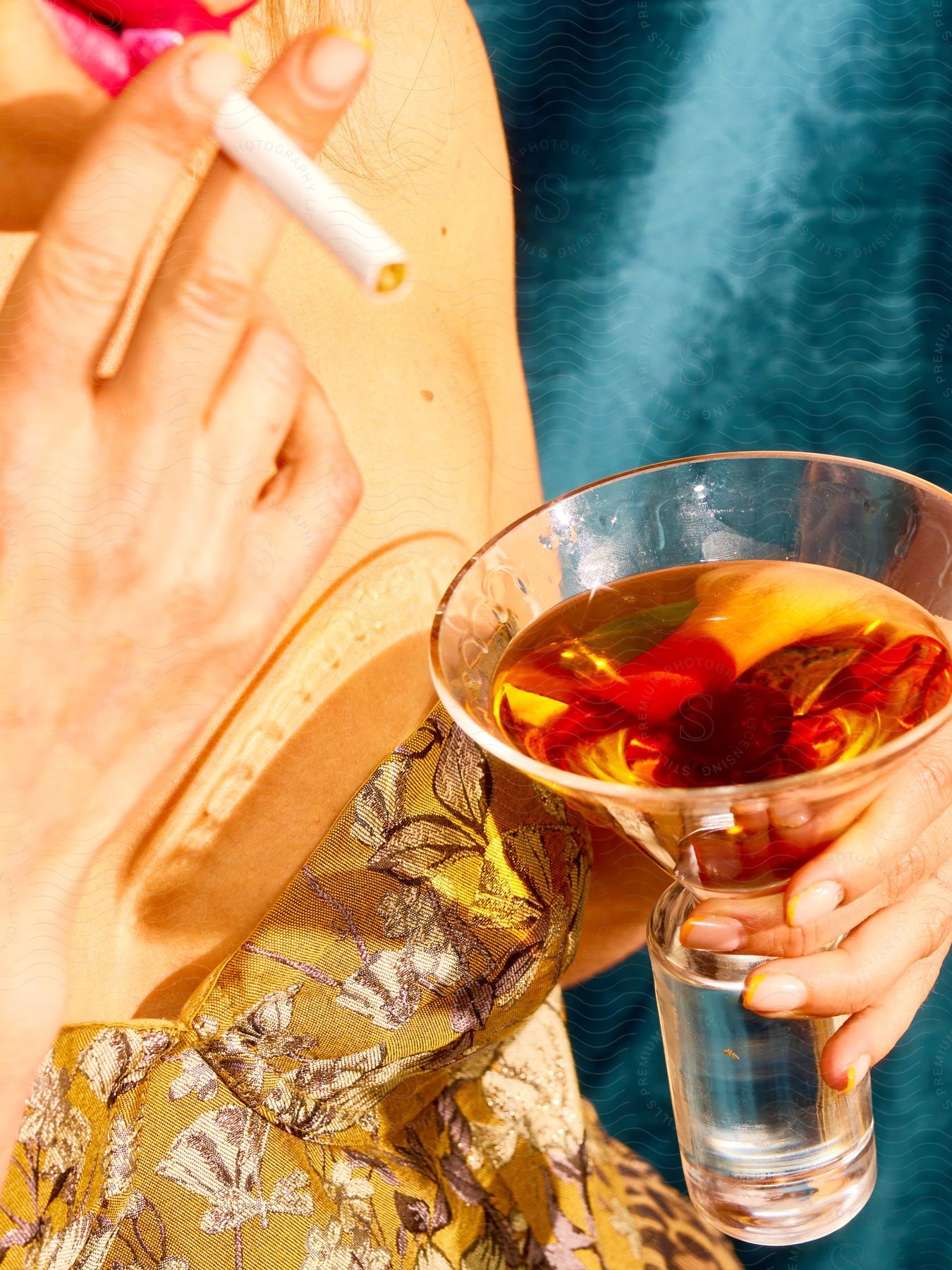 Woman hands holding a glass of olive drink and smoking a cigarette all against a blue background.