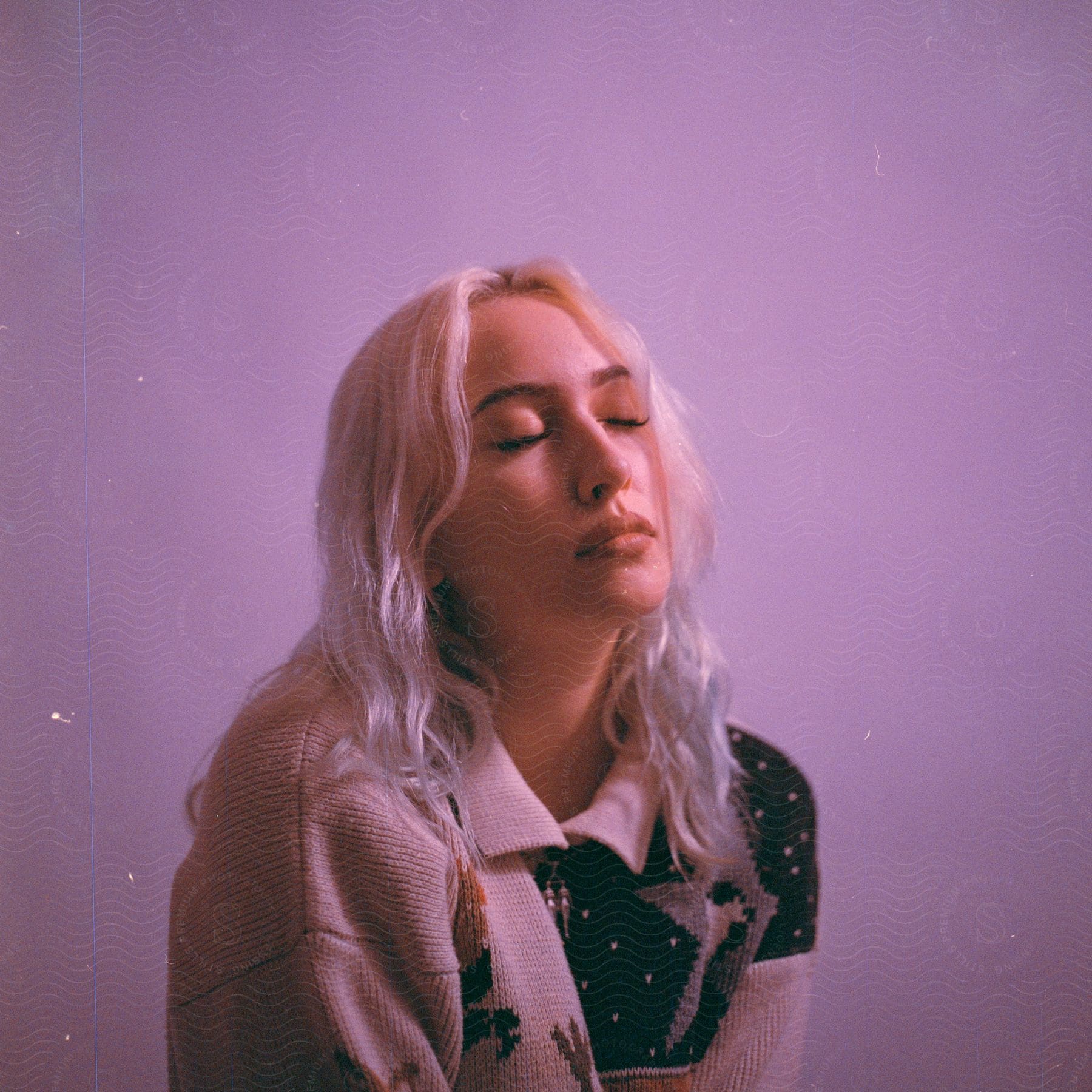 Woman with white hair and a gray sweater with black patterns stands with eyes closed against a pink background