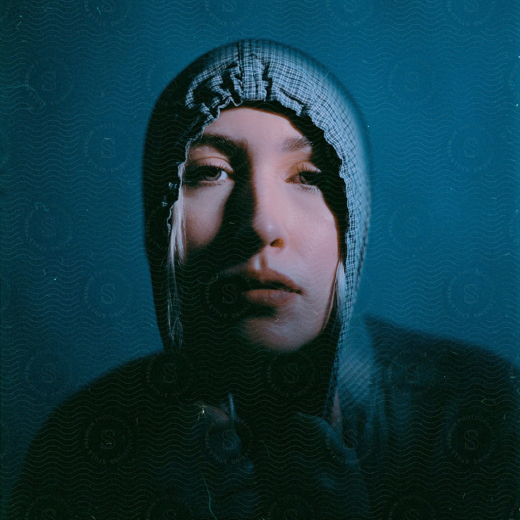 A female wearing a blue hoodie who looks sad under light and dark lighting and a blue background.