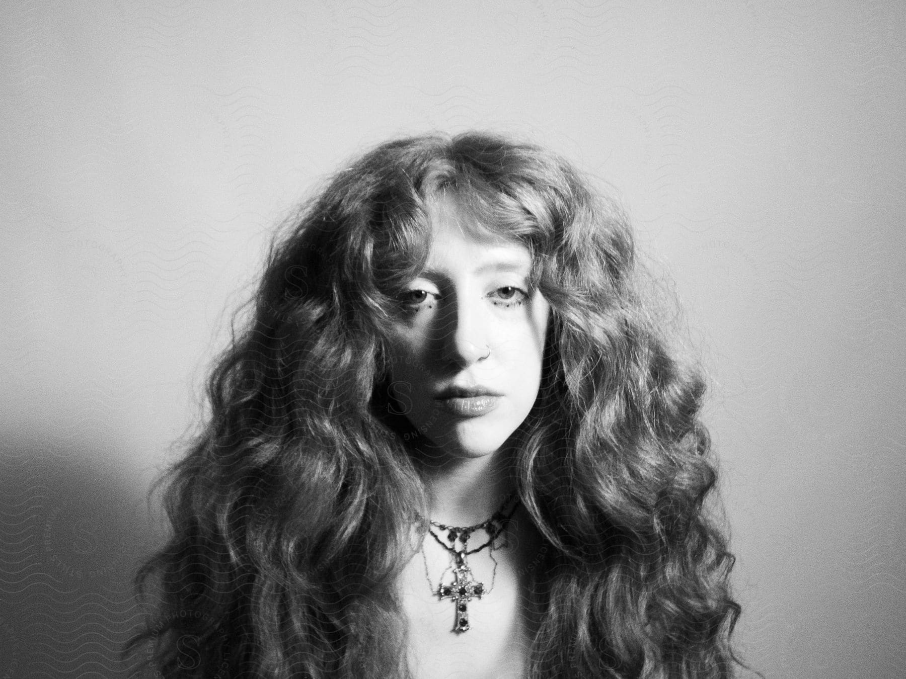 The face of a woman who is posing looking forward with big wavy hair and she is wearing a necklace with a cross and is in black and white tones