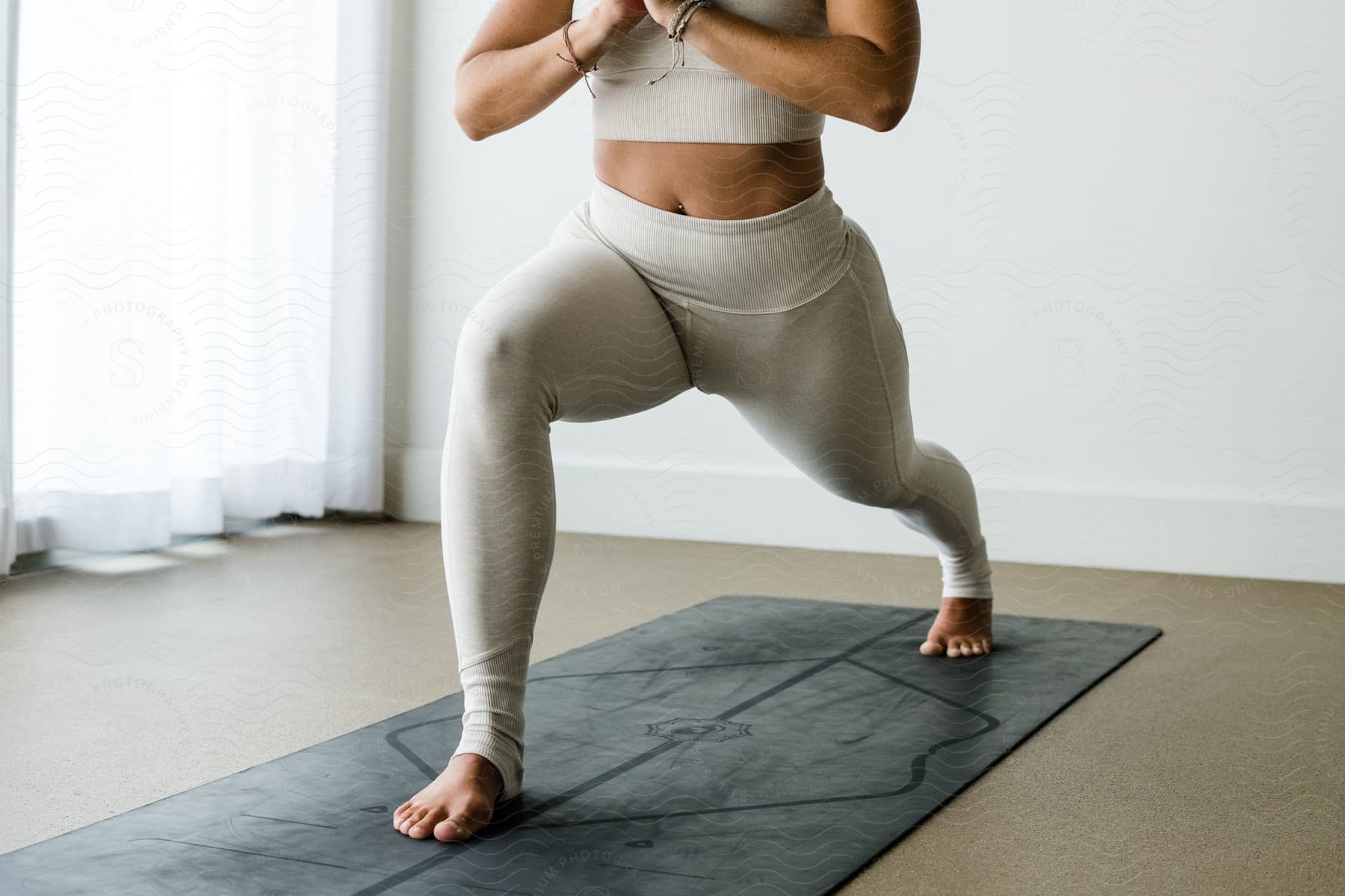 Woman in a yoga pose with hands in prayer position, wearing beige activewear, on a mat in a brightly lit room.