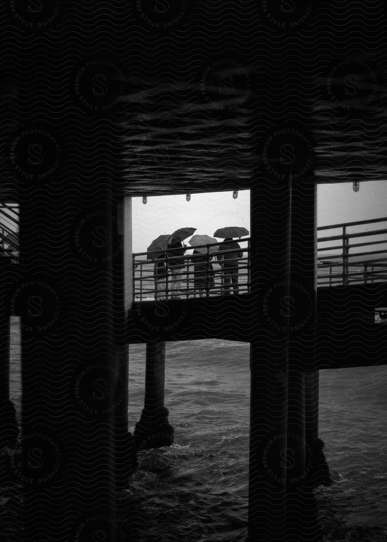 Stock photo of view under pier support pilings with silhouettes of people using umbrellas in the background enjoying the sea.