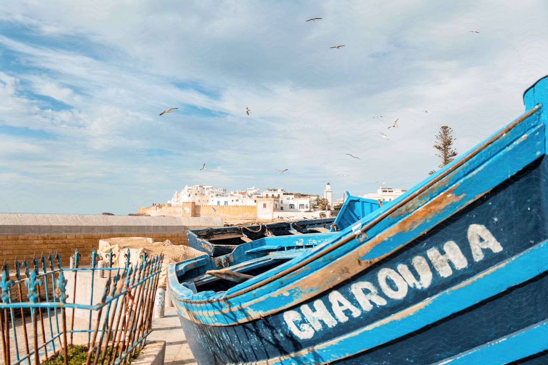 Blue and wooden fishing boat anchored on land with a blue sky with seagulls flying and a coastal town in the background.