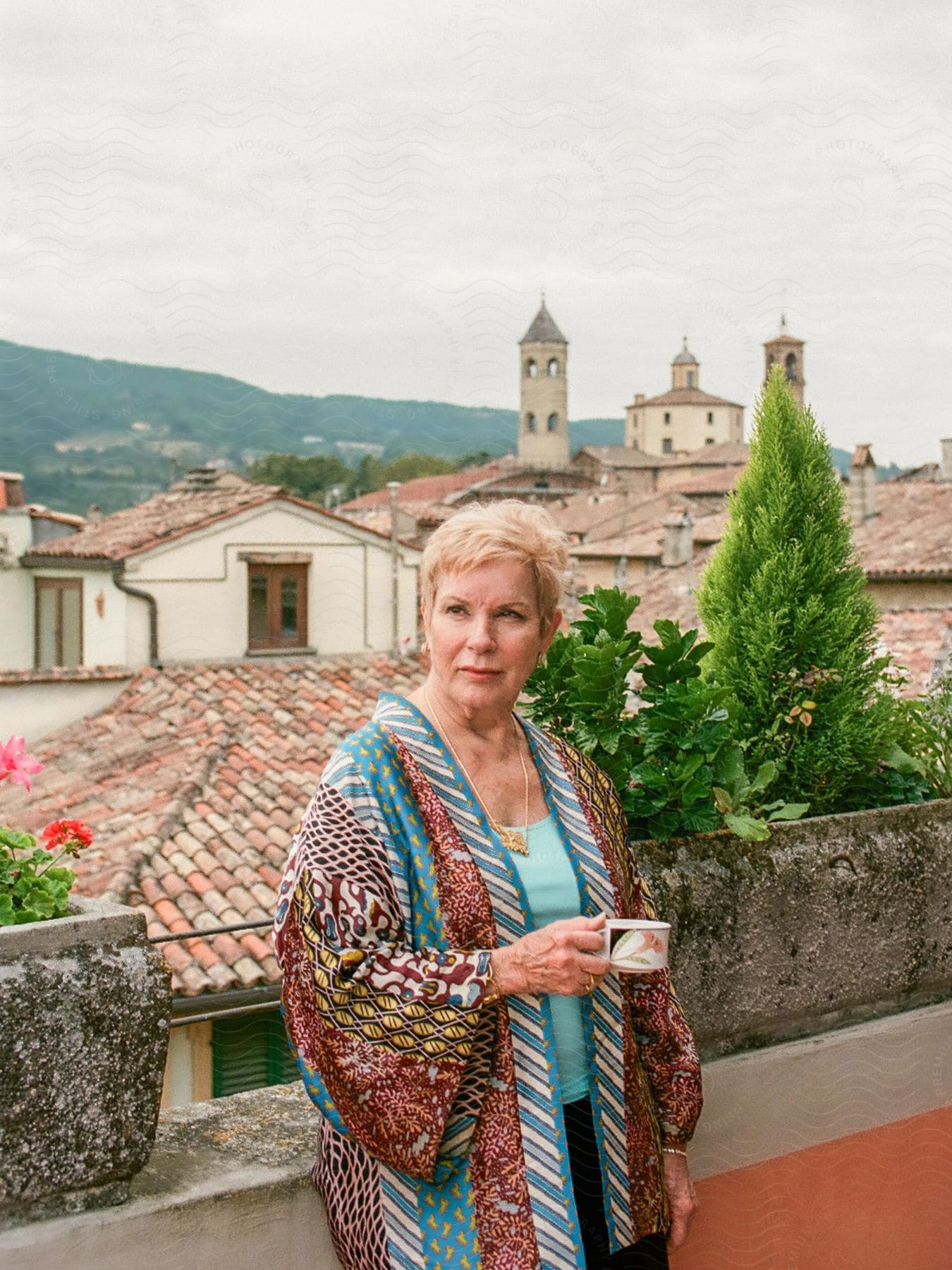 A lady holding a cup in front of a city of roofs and towers of an ancient city.