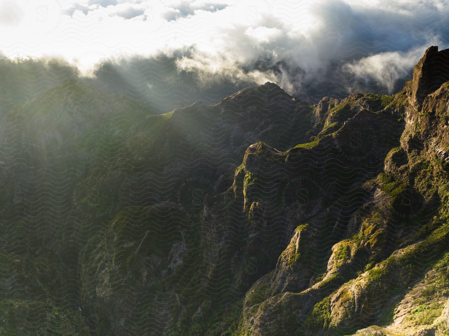 A majestic view of rugged mountains partially shrouded in clouds, illuminated by the sun's rays.