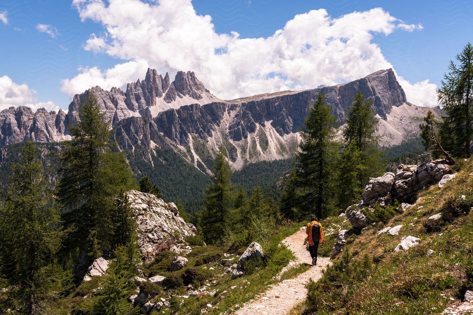 A hiker walks along a gravel trail near a mountain range on a partly cloudy day.