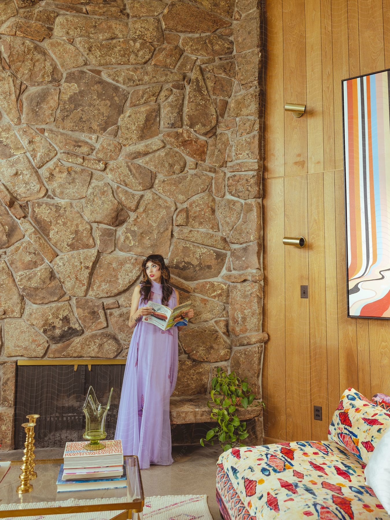 Woman wearing a long lavender gown stands near a fireplace and stone wall holding a book