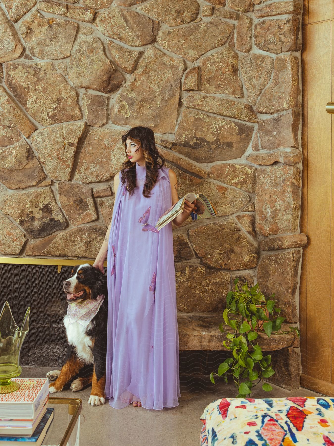 A woman in a long dress, standing next to a dog, holding a book, with a stone wall in the background.